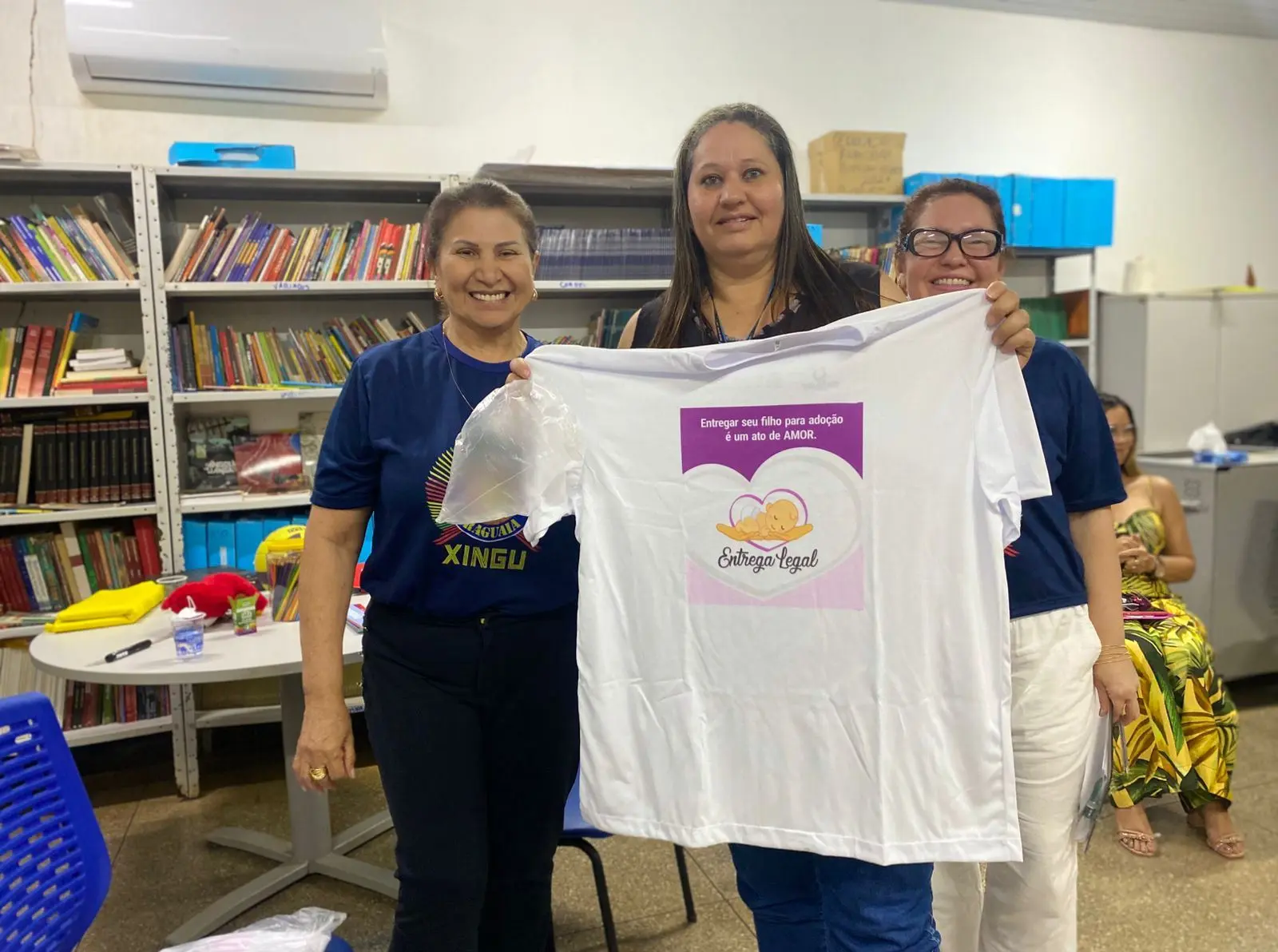 Sala com estantes de livros; três pessoas, uma segura camiseta com mensagem sobre adoção legal. Ambiente acolhedor, educativo, com mesas e cadeiras.