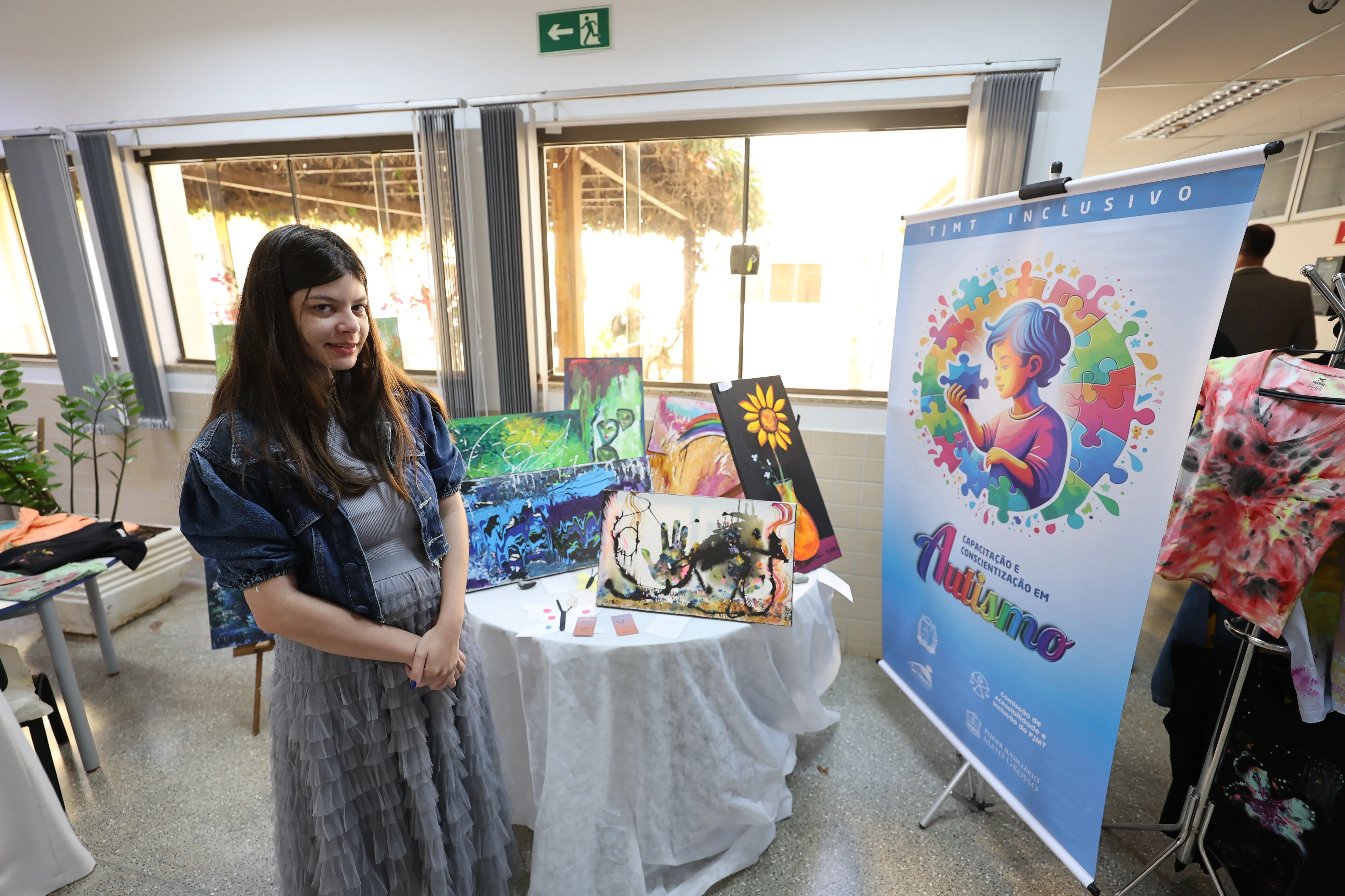 Em espaço claro e bem decorado, jovem posa sorridente ao lado de suas pinturas expostas no TJMT Inclusivo. O ambiente destaca cores vibrantes, natureza e arte como símbolos de sensibilidade e talento.