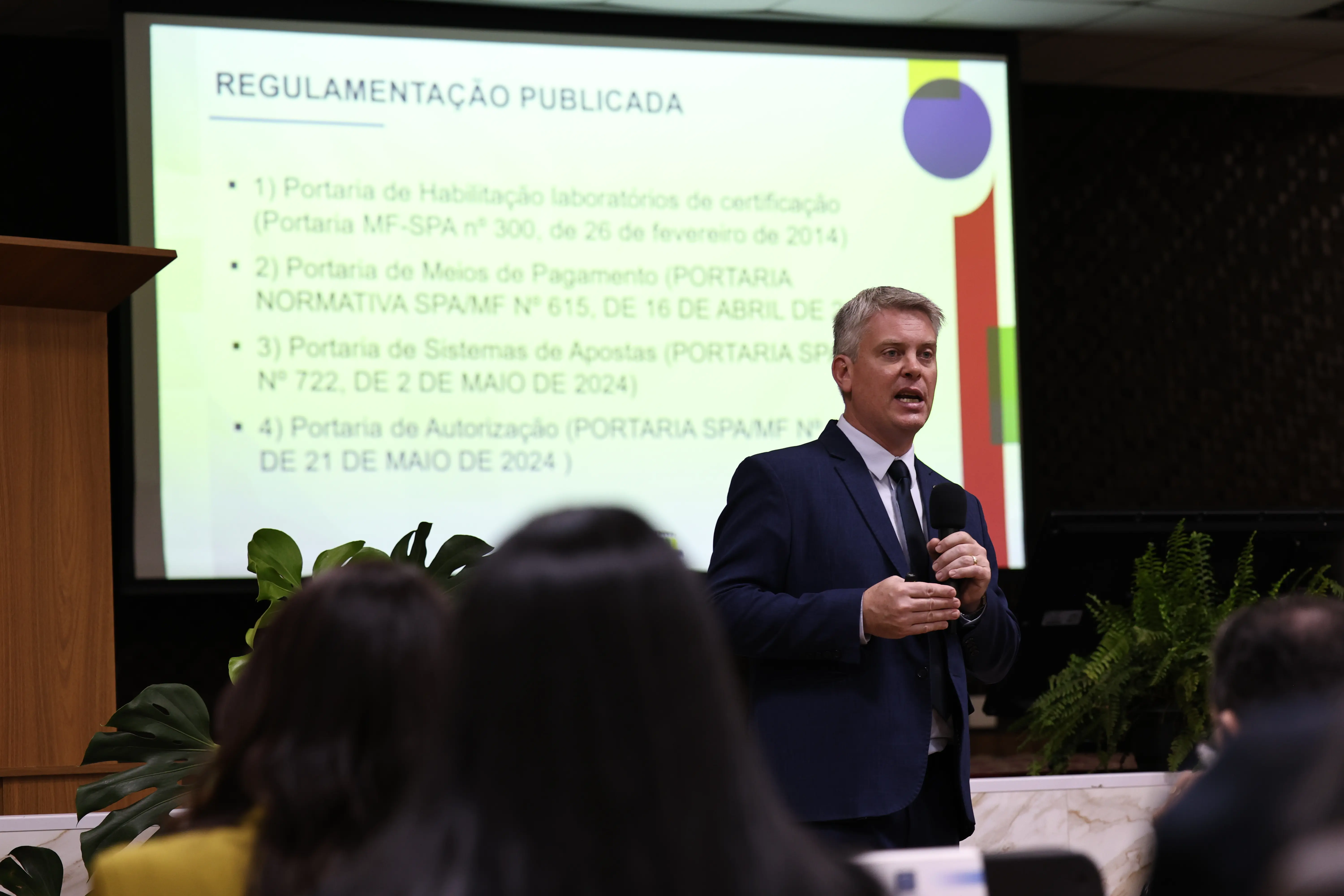 Fábio Macorin, de terno, ministra palestra. Um slide no telão exibe o título "Regulamentação Publicada" e uma lista de portarias.