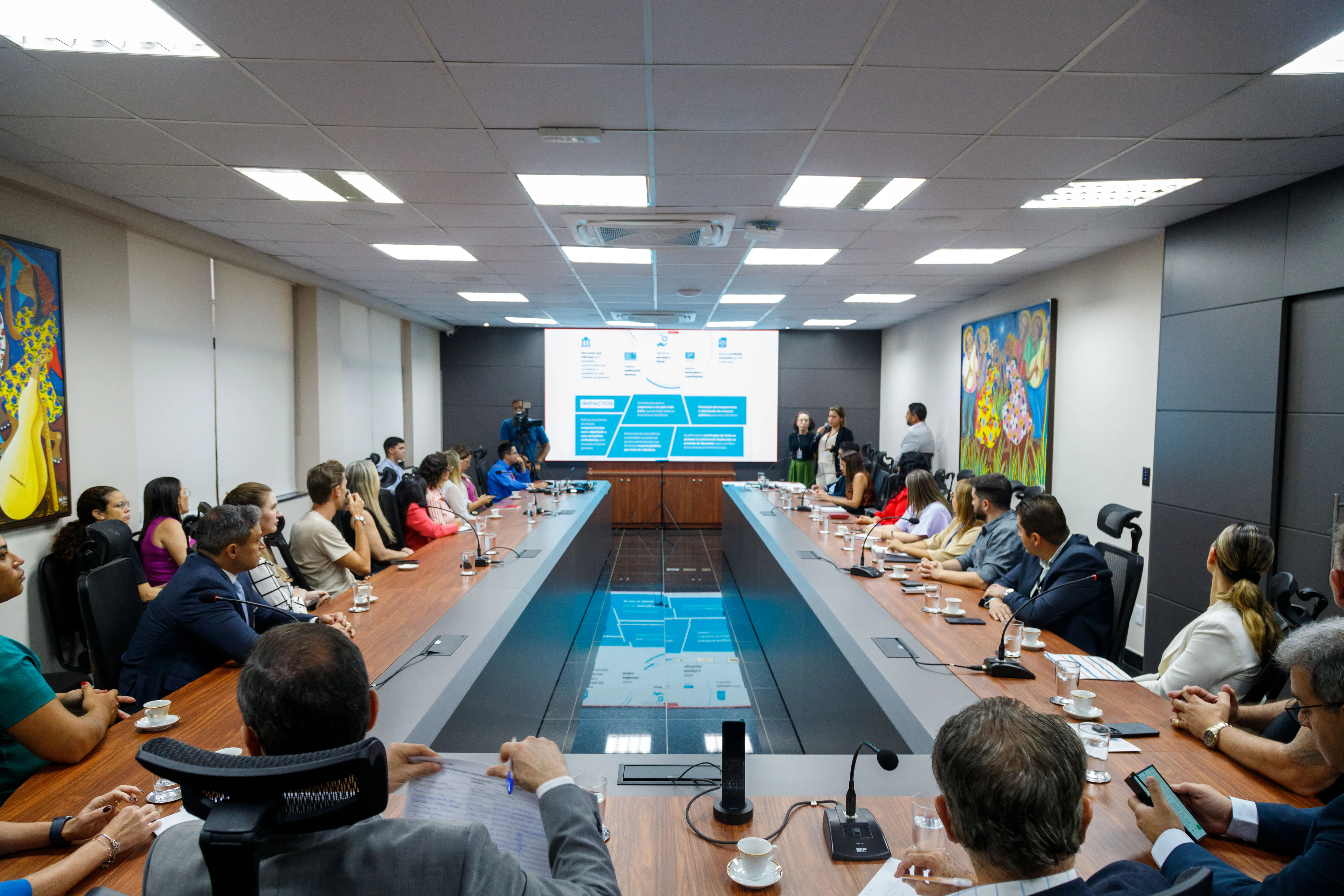 Foto horizontal colorida da sala de reuniões. Uma mesa marrom extensa em formato de U está em destaque com os participantes. Ao fundo da imagem está o telão com a apresentação