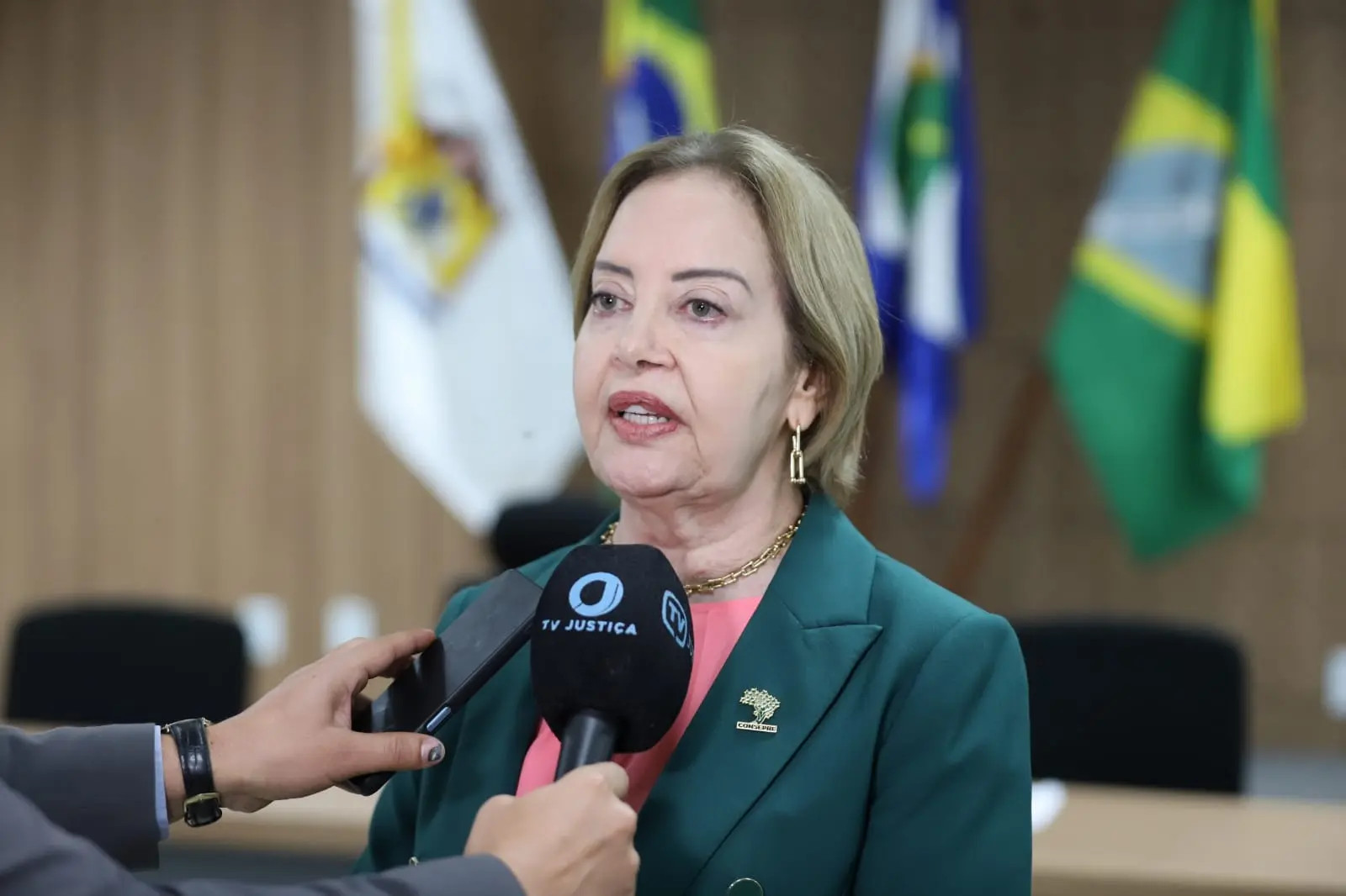 Presidente Clarice concede entrevista. Na foto aparece o microfone preto. Ela é uma mulher branca, de cabelos loiros e curtos