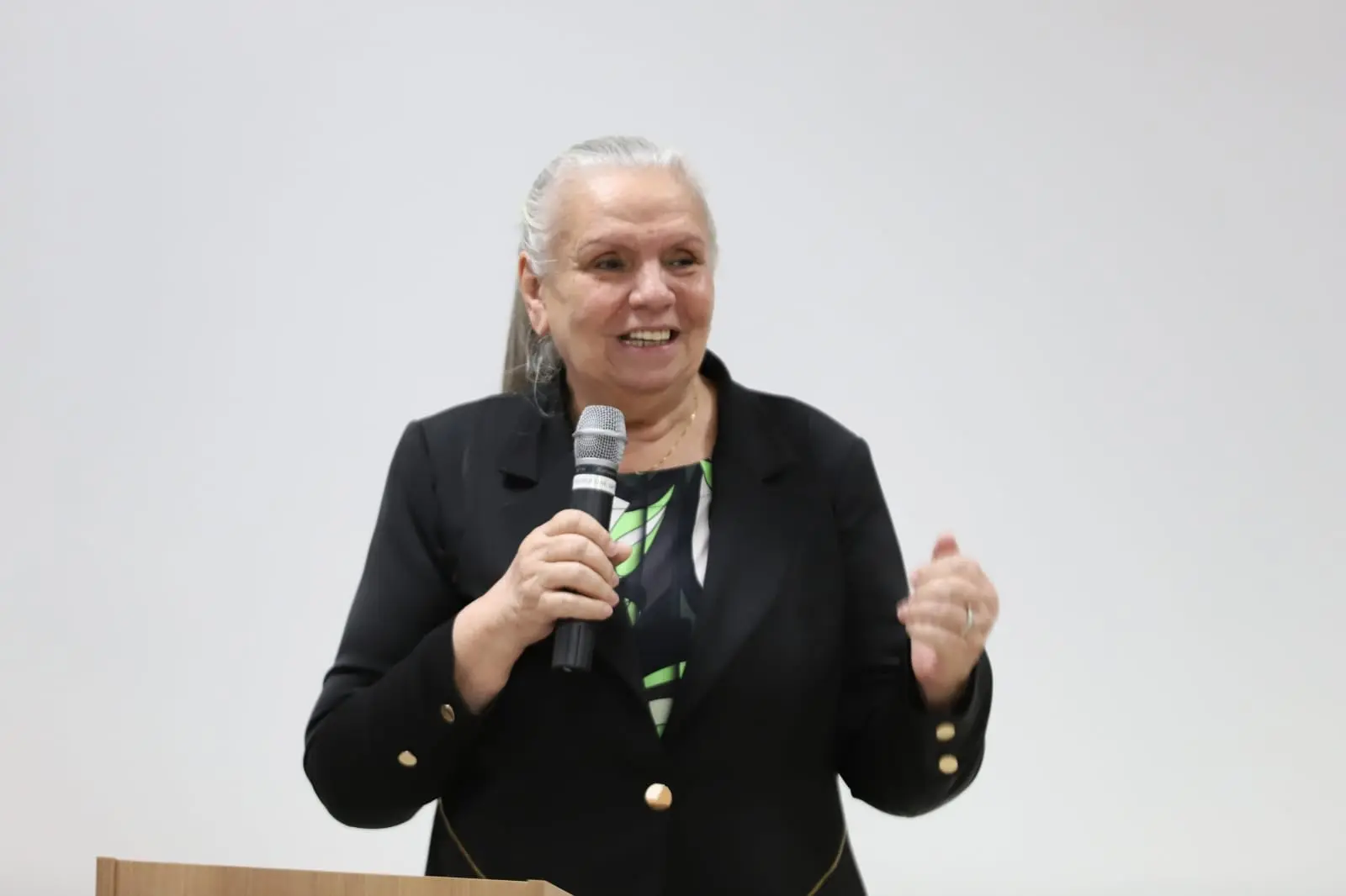 desembargadora Maria Erotides em foto colorida com ângulo fechado. Ela é Uma mulher branca, de cabelos compridos lisos e loiros que estão presos. Usa terno preto e uma blusa preta com estampas verdes. Ela fala ao microfone para os presentes
