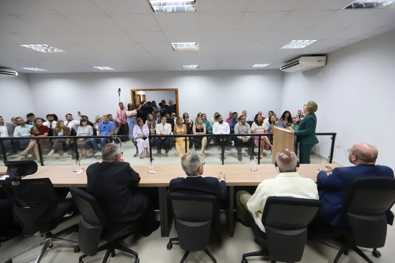Imagem colorida em plano aberto da sala do Tribunal de Juri em Alto Garças. A presidente Clarice Claudino está discursando atrás de um púlpito.
