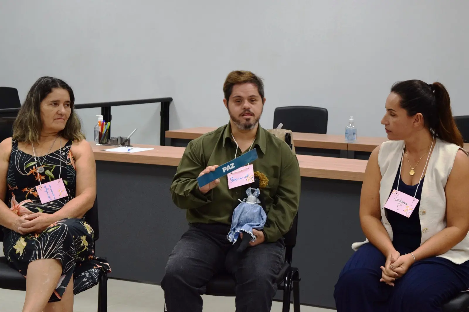Um dos participantes do círculo de paz, que tem síndrome de down segura um boneco com a mão esquerda. Com a mão direita ele segura uma tarja de papel escrito paz