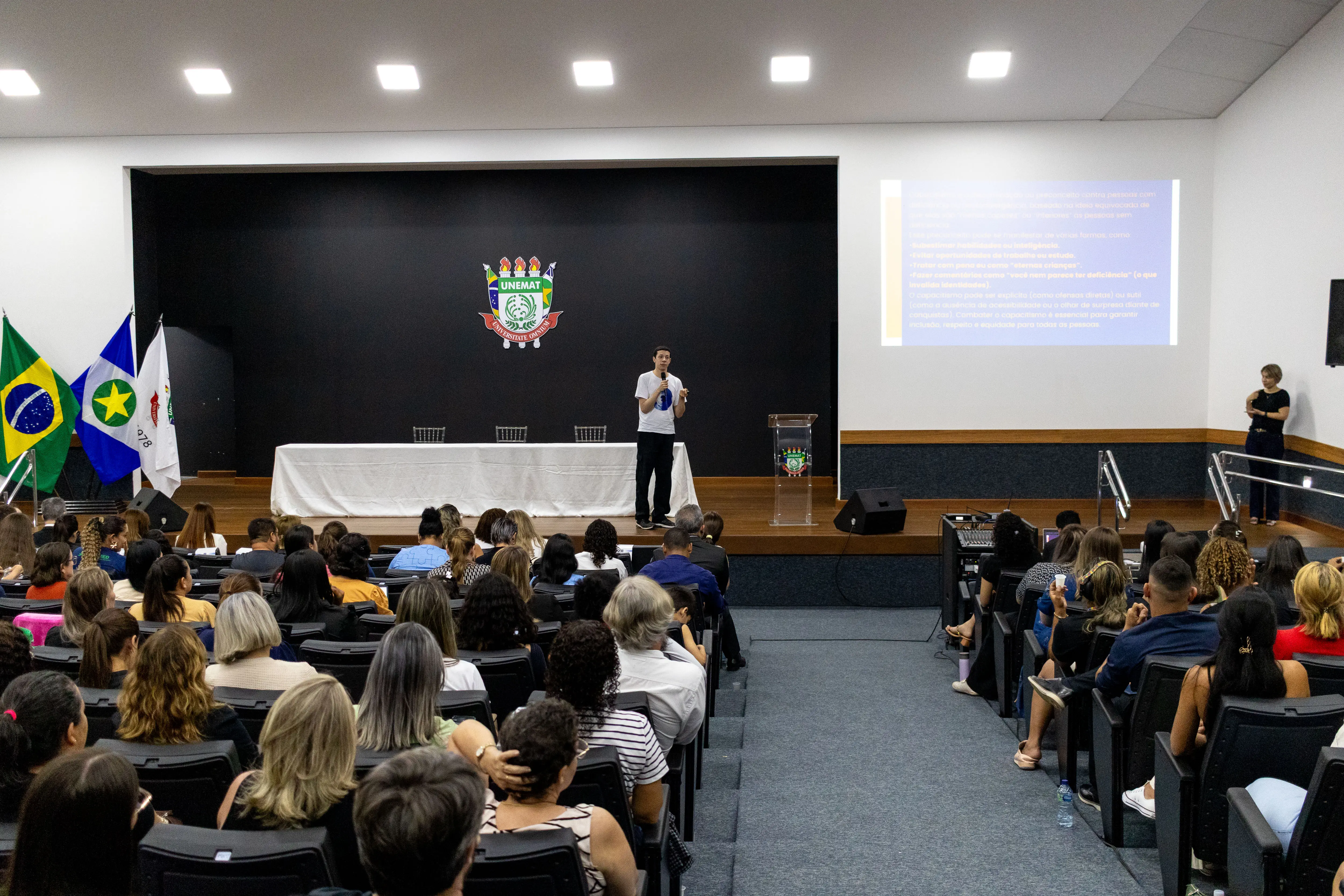 foto em ângulo aberto do auditório da Unemat em Sinop, onde ocorreu o TJMT iNc lusivo. No palco está Nicolas e de frente apra ele, muitas pessoas sentadas em poltronas