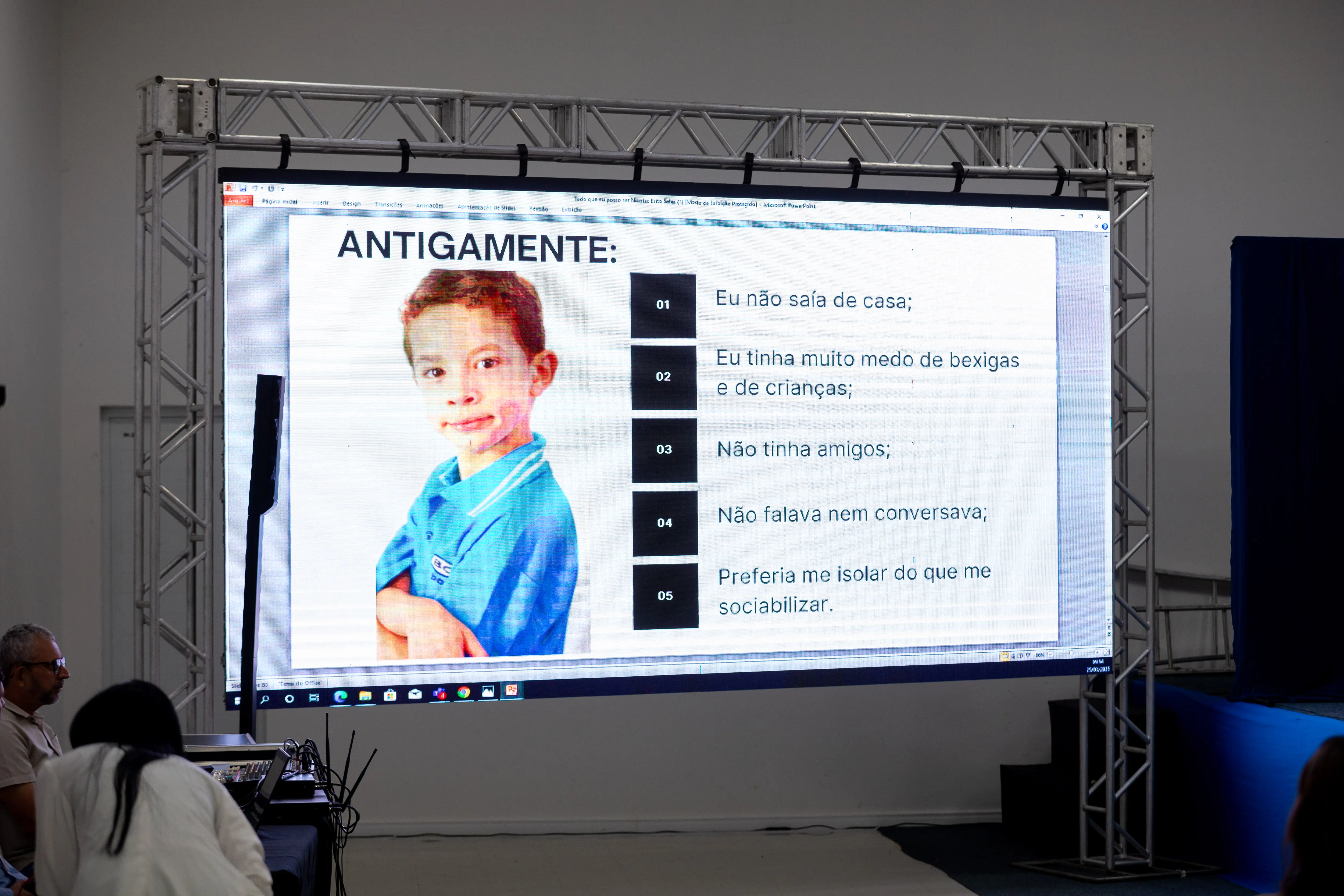 Foto em ângulo fechado de um telão durante o TJMT Inlcusivo em Sorriso. A imagem no telão mostra slide de um menin o escrito: antigamente seguido de tópicos que mostram com o Nicolas, autista, se com portava antes