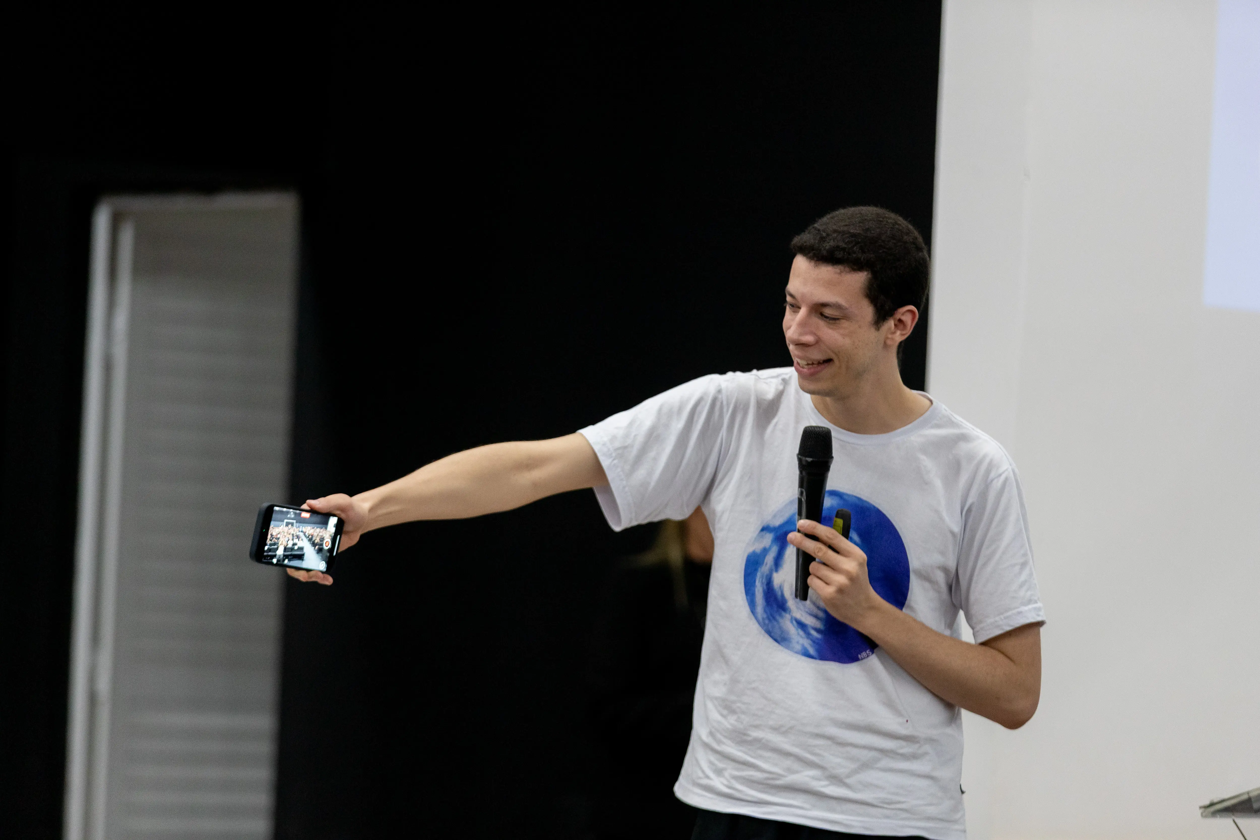 Nicolas durante palestra do TJMT Inlcusivo, em Sinop. Ele é um rapaz magro, branco, usa camiseta branca, segura microfone com mão esquerda e um celular com a mão direita