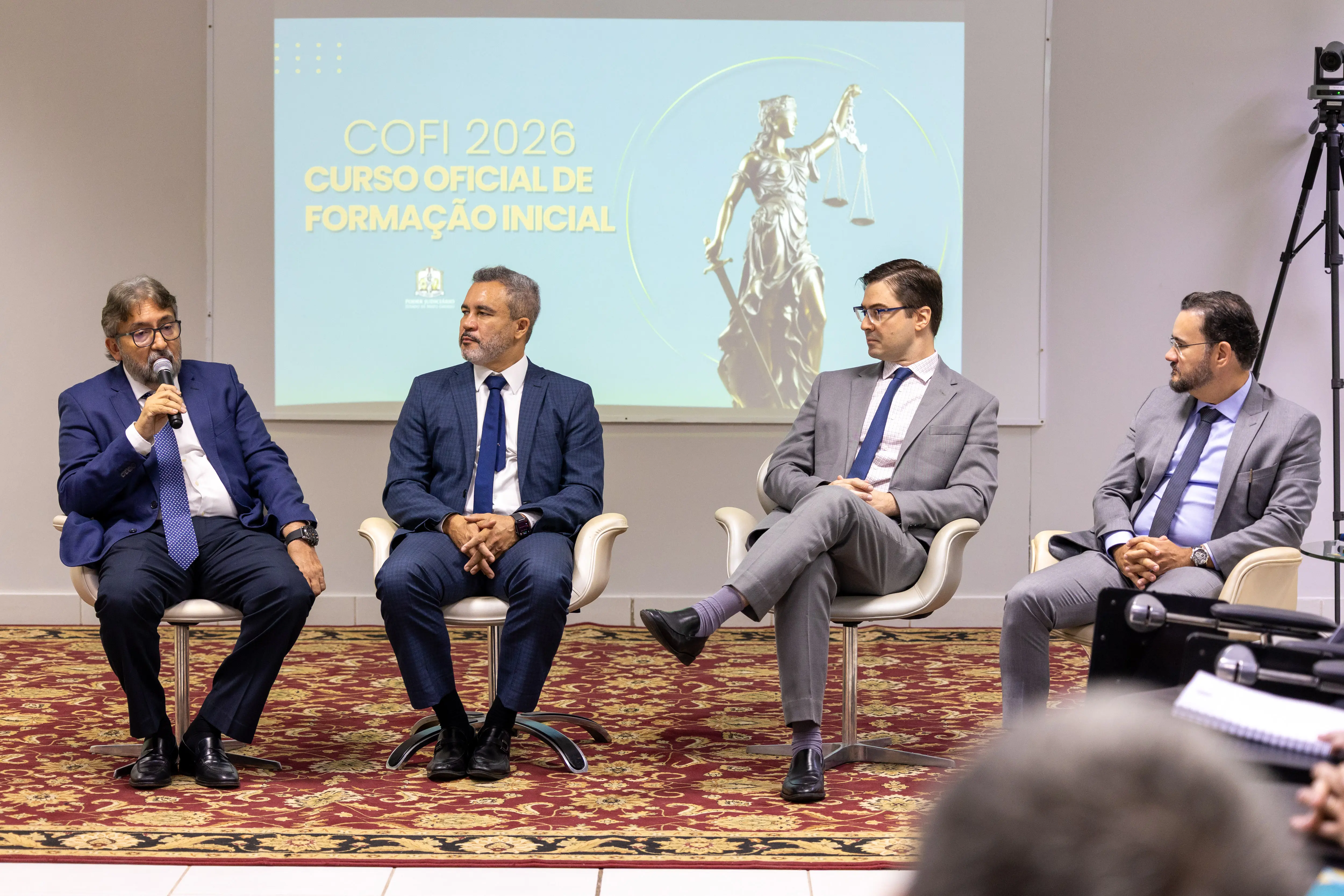 Público acompanha exposição no auditório da ESMAGIS-MT. Ao fundo, banner do COFI 2026 e estrutura de transmissão. O ambiente é organizado, com clima acadêmico e institucional.