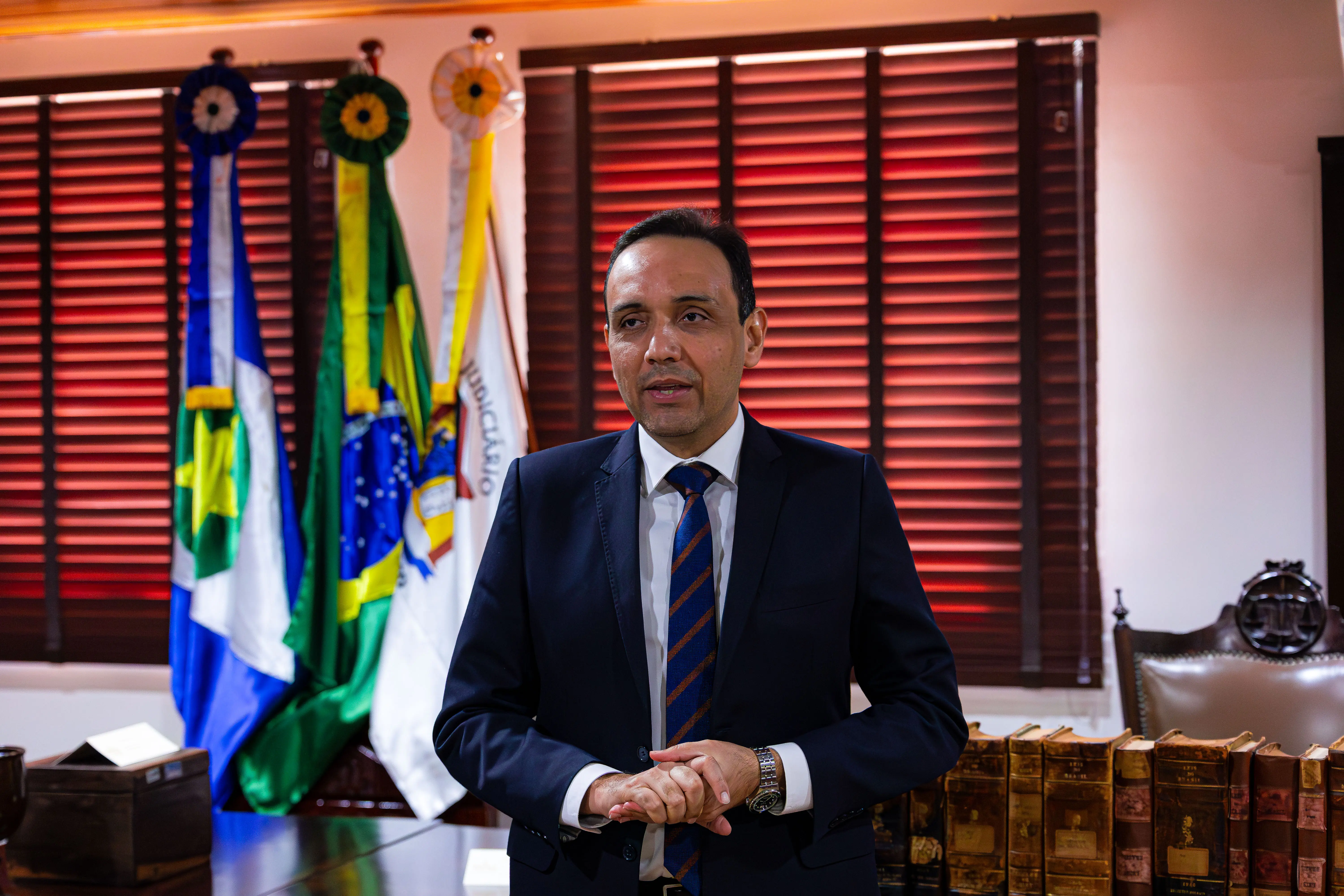 Homem de terno azul-marinho e gravata listrada fala em pé, com bandeiras do Brasil e de Mato Grosso e o brasão do Judiciário ao fundo.