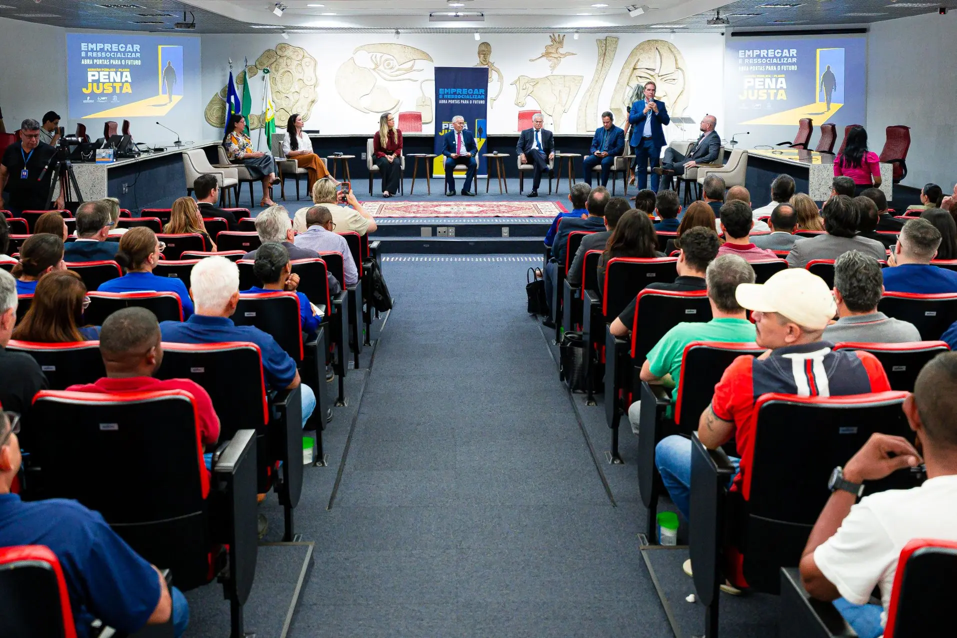 Foto horizontal em plano aberto que mostra o plenário do TRT-MT lotado de pessoas na plateia (que está de costas na foto). No palco, autoridades estão sentadas em poltronas, durante audiência pública.