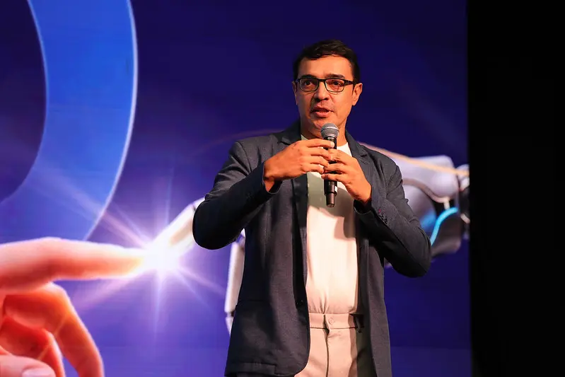 Foto horizontal que mostra Welkey Costa da cintura pra cima, no palco do FestLabs, proferindo palestra. Ele é um homem de pele morena, cabelo castanho escuro, usando calça bege, camiseta branca e blazer cinza escuro.