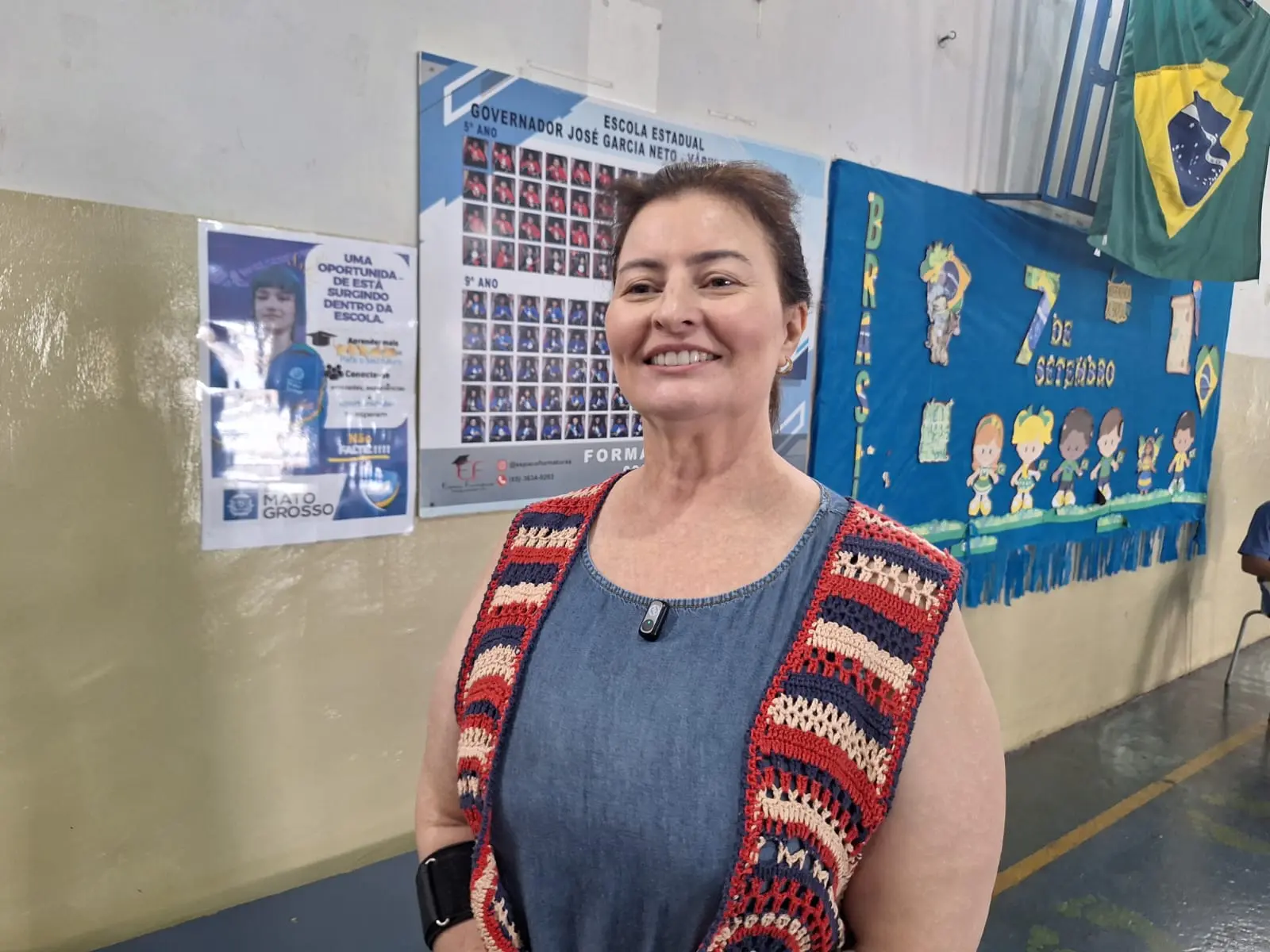 Foto horizontal que mostra a professora Daniela Bueno, da cintura pra cima, em pé no pátio da escola. Ela é uma mulher branca, de cabelos castanhos, lisos e presos, usando vestido jeans e colete de crochê nas cores azul, vermelho e bege. Atrás dela aparece uma parece com mural e bandeira do Brasil.