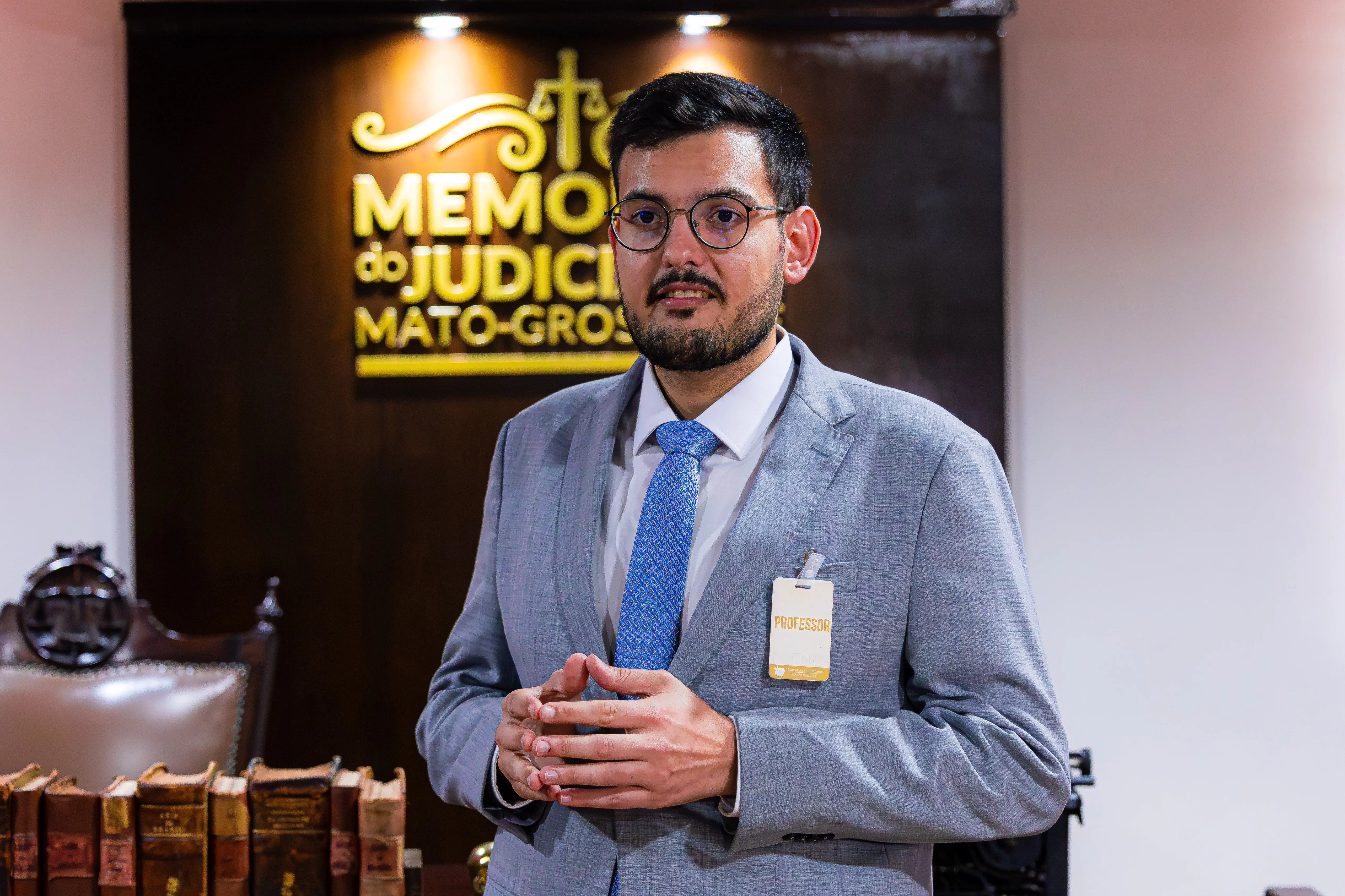 Homem de óculos, barba e terno cinza, com crachá "PROFESSOR", fala em pé no Memorial do Judiciário, tendo o letreiro da instituição e livros antigos ao fundo.
