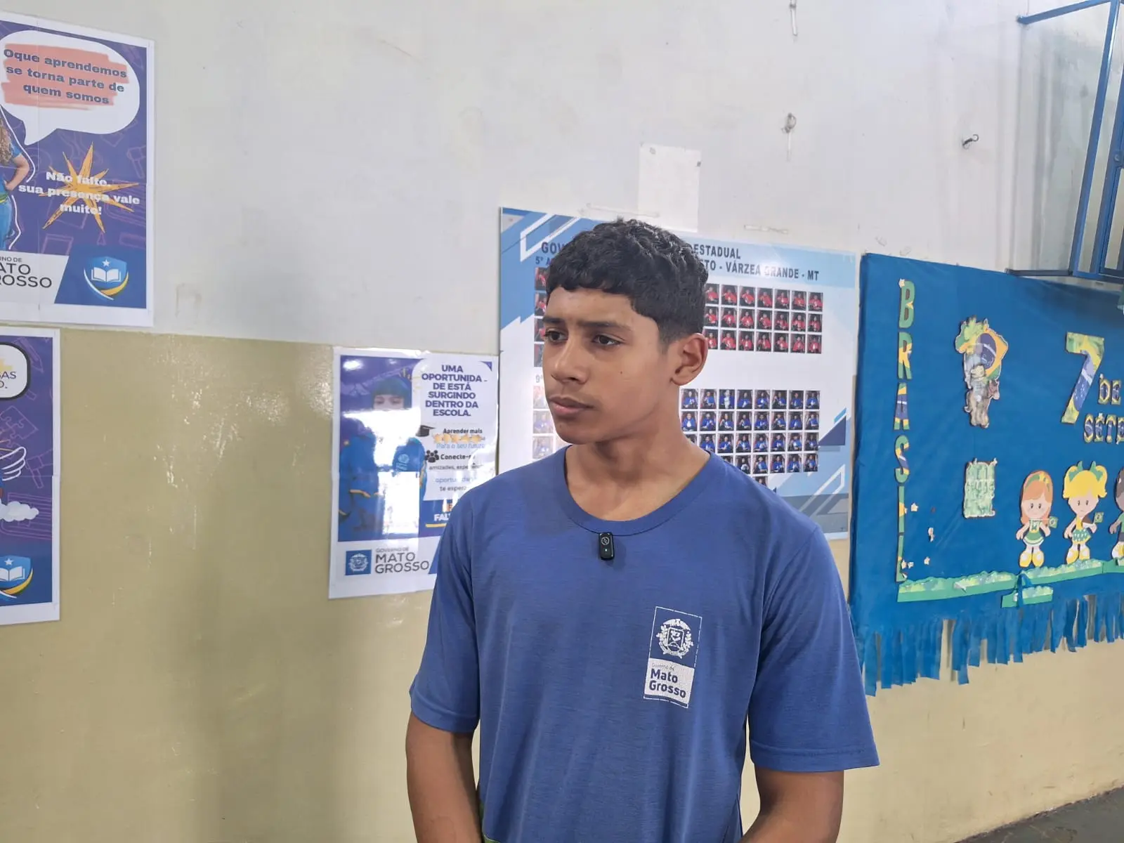 Foto horizontal que mostra o estudante Ezequiel Teixeira em pé, no pátio da escola. Ele é um adolescente negro, de olhos e cabelos castanho escuro, usando uniforme escolar azul. Atrás dele, a parede tem placas de formandos, mural e cartazes.
