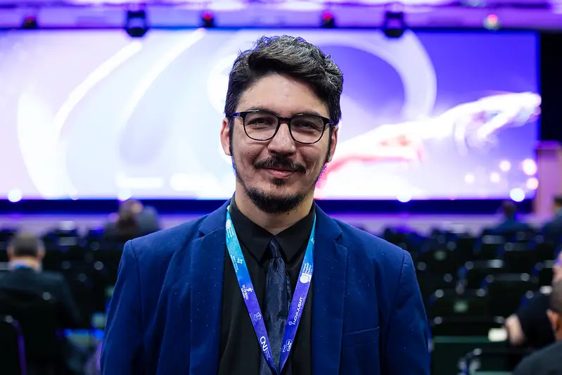 Foto horizontal que mostra Dimmy Magalhães, participante do FestLabs Cuiabá, posando para a foto. Ele é um homem branco, de cabelo, olhos e barba castanho escuro, usando camisa preta, terno azul, óculos de grau e crachá do evento. Ao fundo, salão de eventos com palco iluminado em tons de azul e roxo