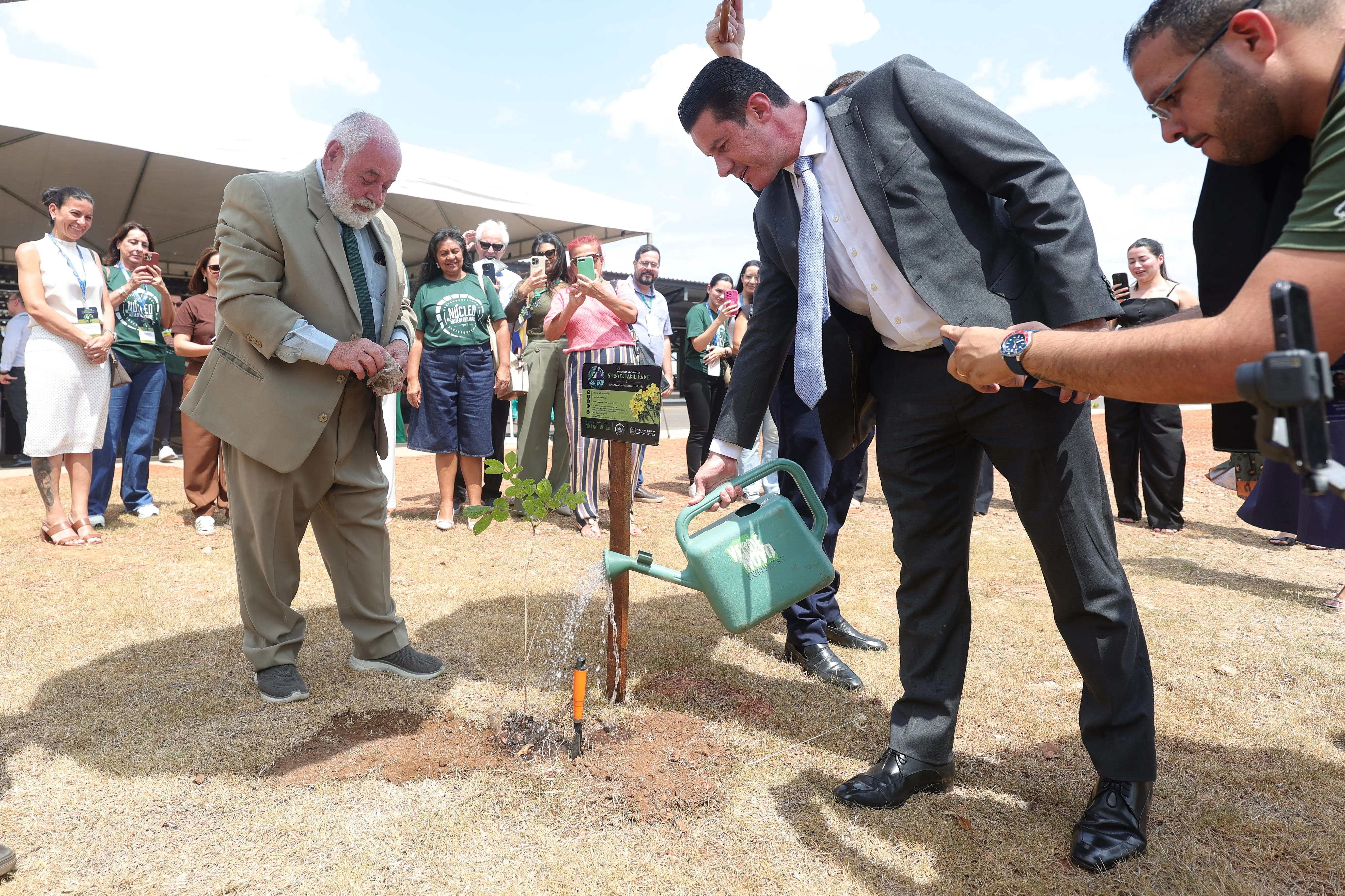 Foto horizontal em plano aberto que mostra os desembargadores José Zuquim e Rodrigo Curvo em volta de uma muda plantada por Zuquim e regada por Curvo. Várias pessoas aparecem ao redor, observando e fotografando. O céu aparece ensolarado e o solo com grama seca.