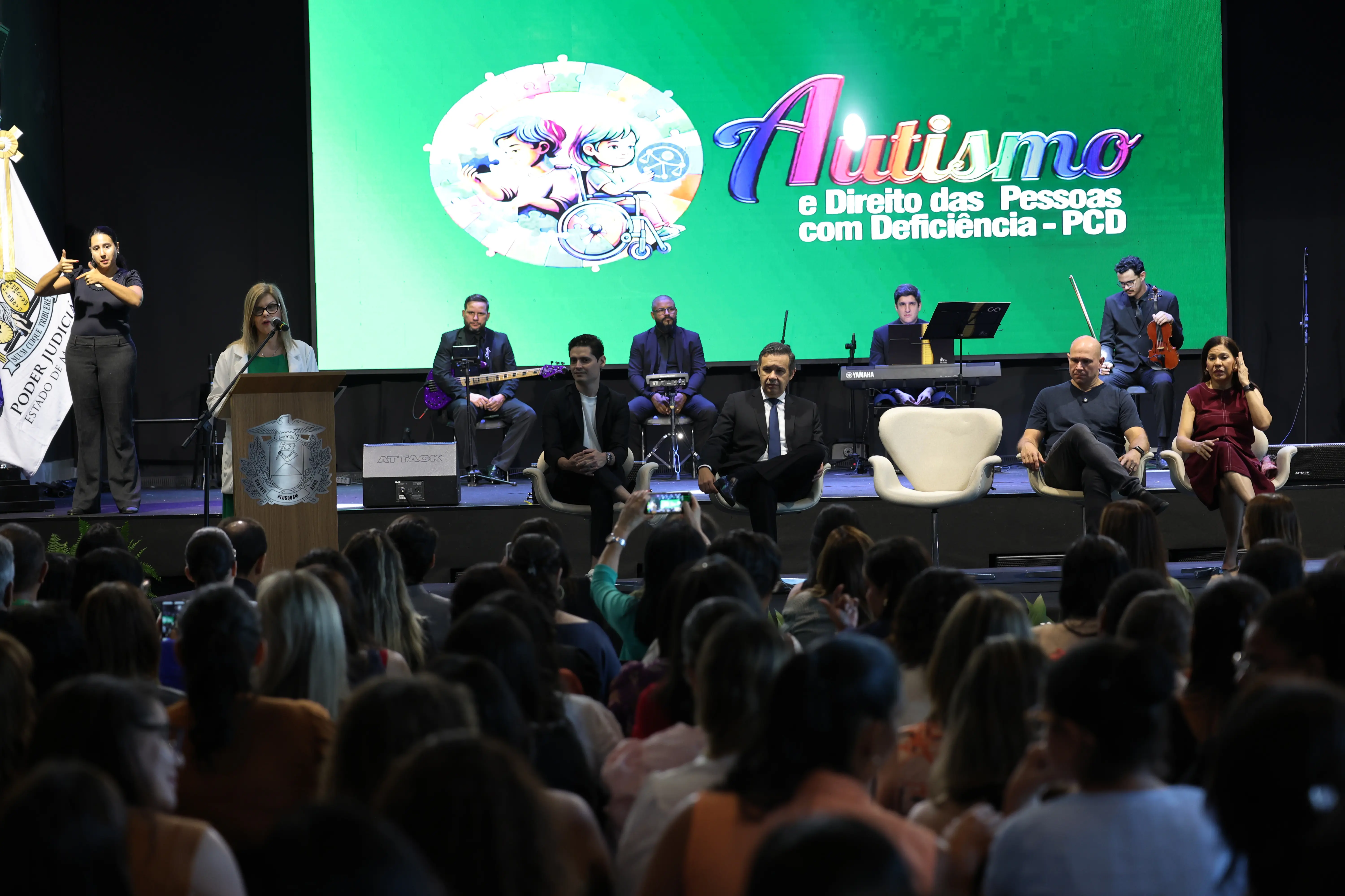 Evento em auditório com palco e telão verde escrito "Autismo e Direito das Pessoas com Deficiência - PCD". No palco, autoridades, músicos e intérprete de Libras. Plateia atenta em primeiro plano.