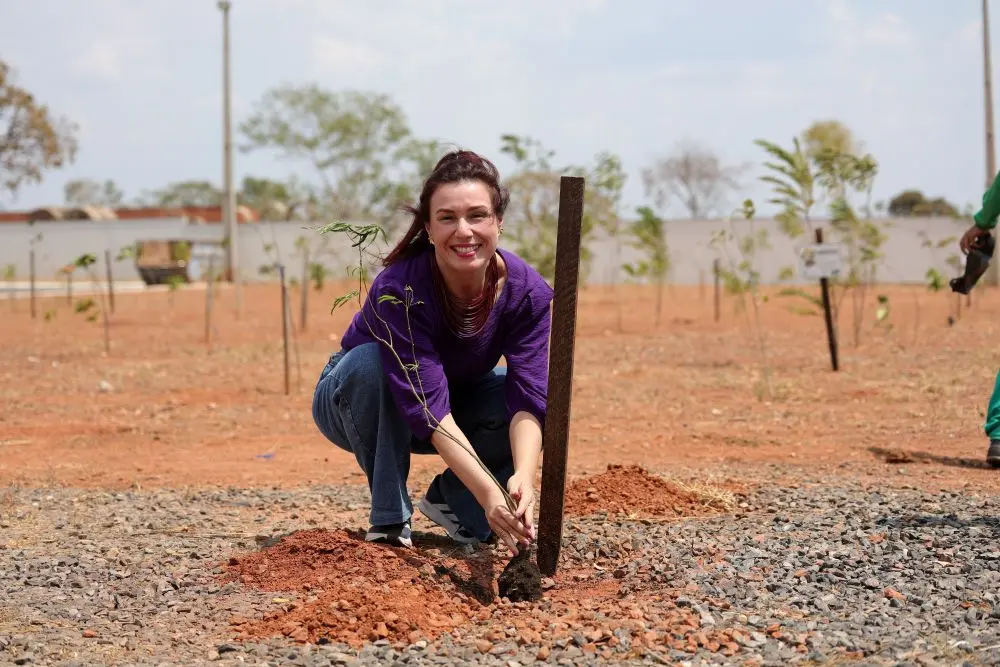 Foto horizontal em plano aberto que mostra a servidora do TJ de Rondônia, Samira Alvim, agachada ao solo, plantando uma muda de árvore. Ela sorri para a foto. Ela é uma mulher branca, ruiva, usando calça jeans e camisa roxa. Atrás dela, aparece um enorme terreno com centenas de mudas plantadas.
