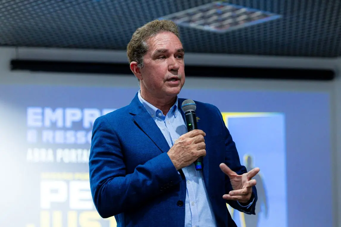 Foto horizontal que mostra o presidente da Fecomércio-MT, Sebastião dos Reis, falando ao microfone em pé, no plenário do TRT-MT. Ele é um homem branco, de cabelos castanhos claros, usando camisa azul clara e terno azul. 
