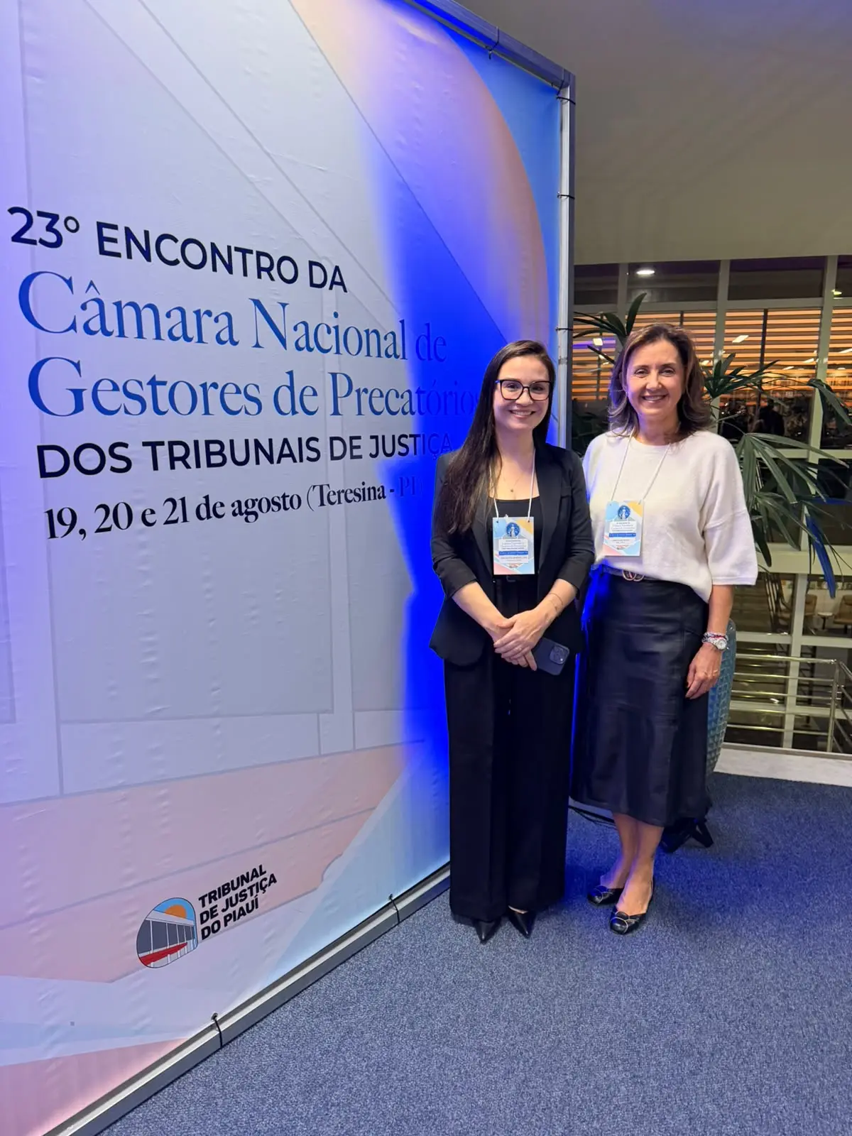 Fotografia em plano inteiro de duas mulheres sorridentes em pé, lado a lado, em um ambiente interno e iluminado. Elas são a juíza-auxiliar da presidência, Christiane Costa Marques Neves, e a servidora da Central de Precatórios, Angelita Dornelles.  Elas estão sobre um carpete azul. À esquerda, um gr