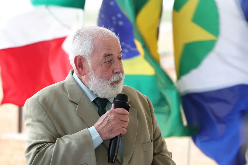 Foto horizontal que mostra o desembargador José Zuquim Nogueira, do busto pra cima, falando ao microfone. ele é um senhor branco, de cabelo e barba brancos, olhos claros, usando camisa e terno em tons de verde. Atrás dele aparecem mastros com bandeiras de Várzea Grande, de Mato Grosso e do Brasil.