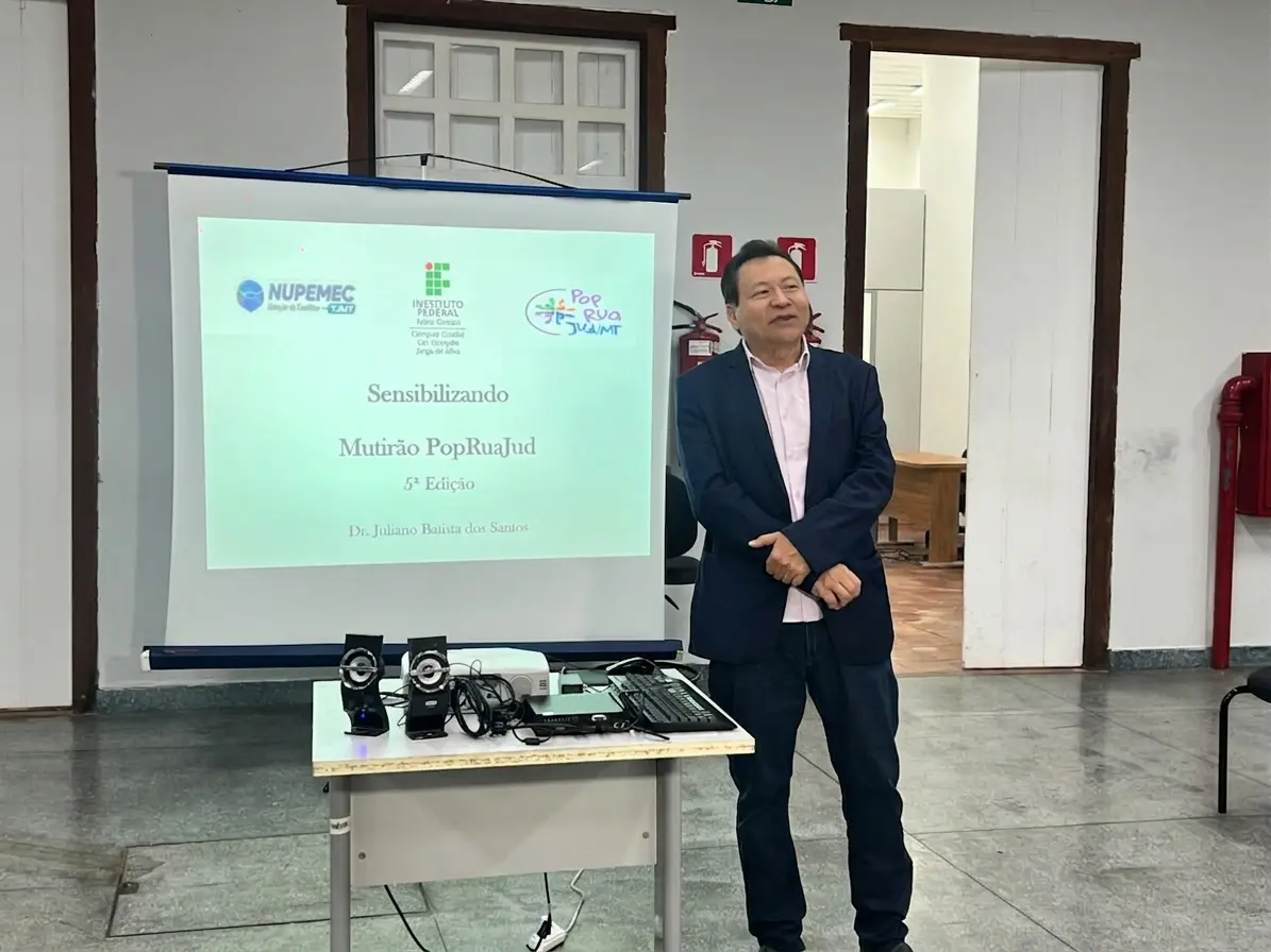 Homem asiático, cabelos grisalhos, camisa social rosa e blazer azul-marinho, discursa em pé ao lado de telão com identificação "Dr. Juliano Batista dos Santos".