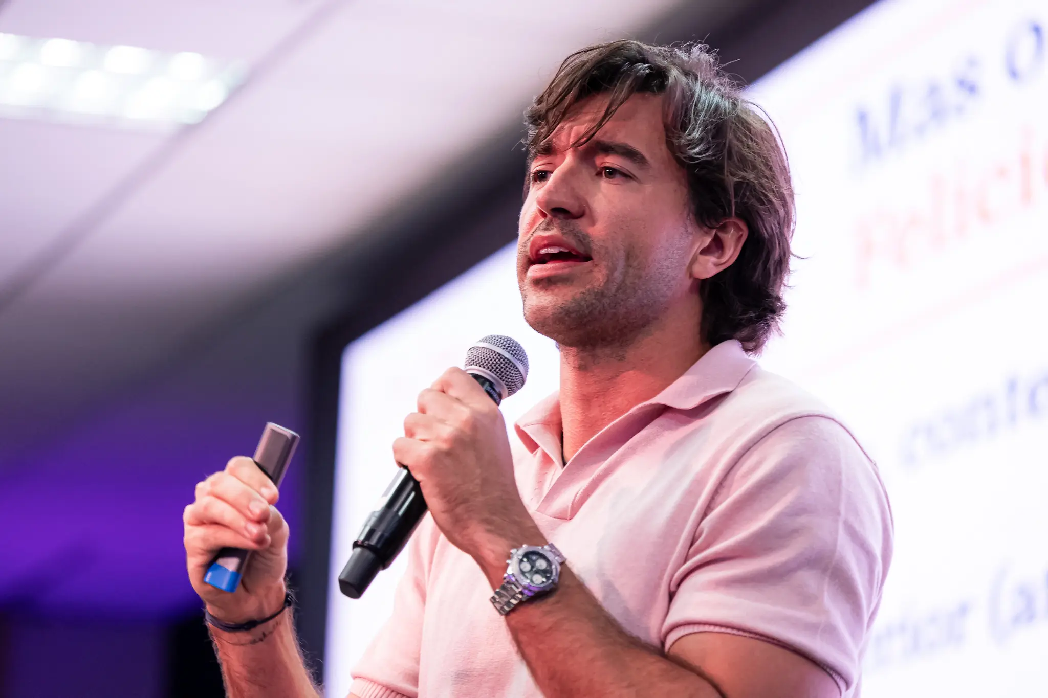 Close de homem de cabelos escuros e camisa polo rosa, segurando microfone e controle de slides, falando em palco durante palestra.