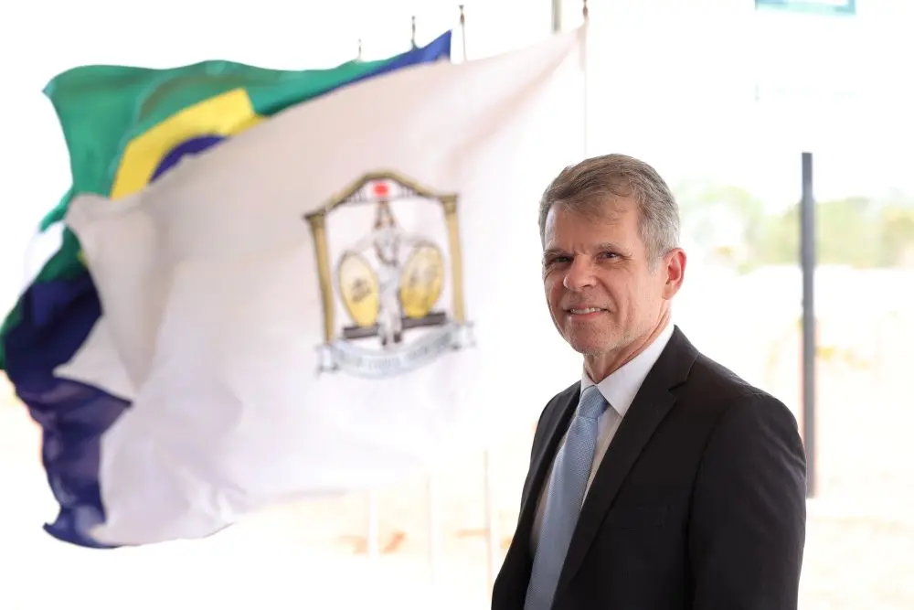 Foto horizontal que mostra o juiz Ângelo Judai Júnior, do busto pra cima, sorrindo para a foto. Ele é um senhor branco, de cabelo grisalho, usando camisa branca, gravata azul clara e terno preto. Atrás dele aparecem bandeiras agitadas pelo vento, com destaque à bandeira do Poder Judiciário.