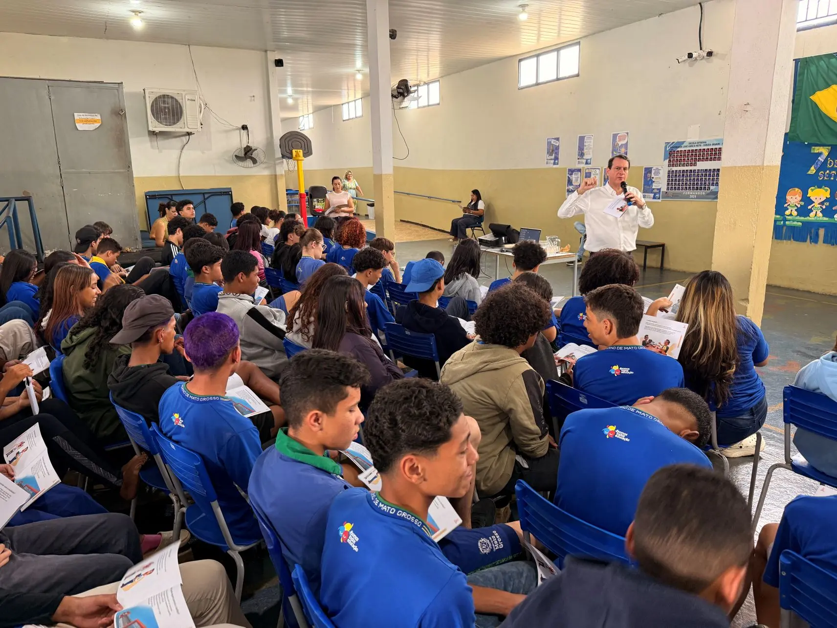 Foto horizontal em plano aberto que mostra dezenas de estudantes com uniforme azul sentados assistindo a uma palestra, no pátio da escola. O palestrante é um homem branco, usando camisa branca e falando ao microfone. 