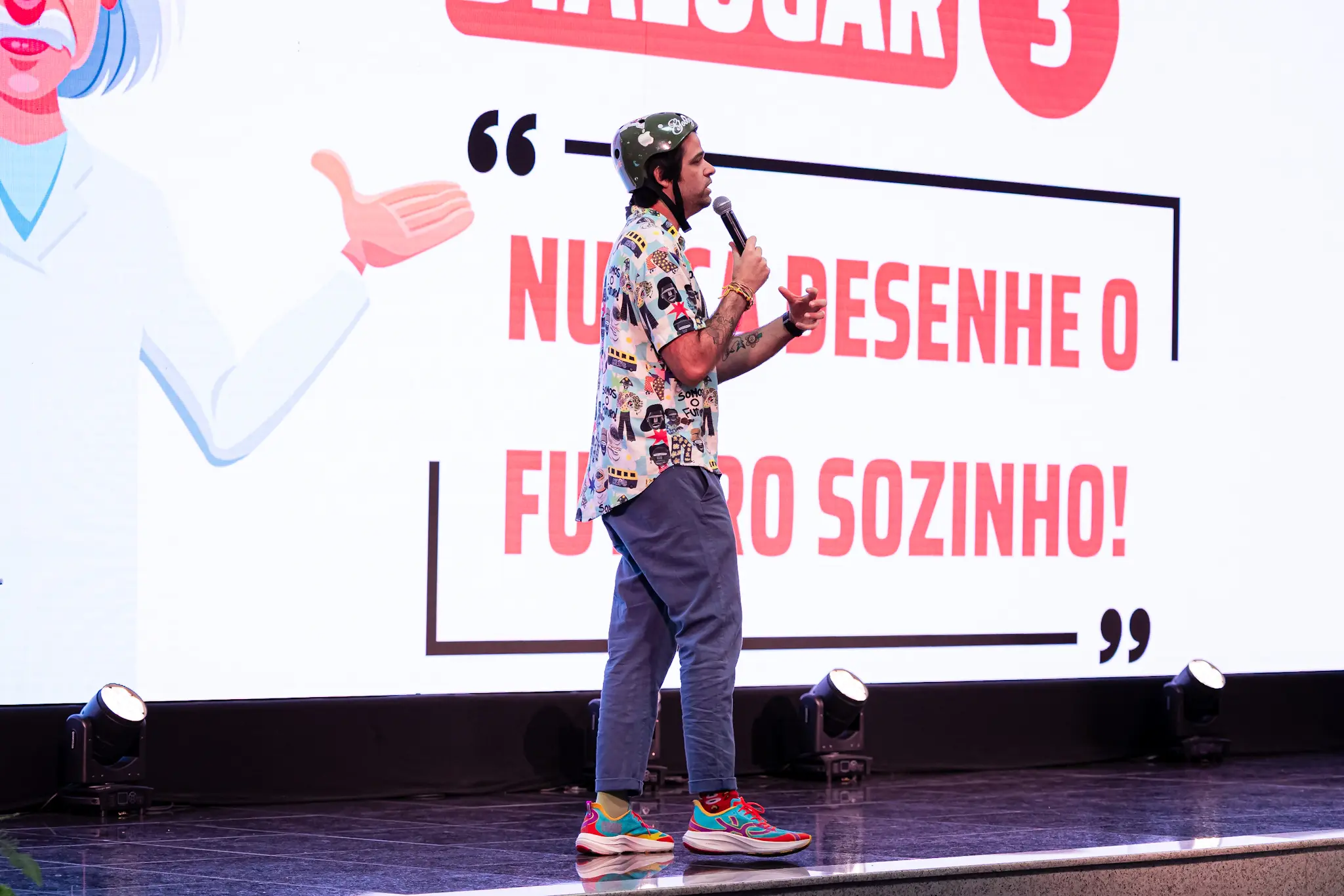 Homem de capacete verde, camisa estampada e tênis coloridos fala ao microfone em um palco. Ao fundo, um telão exibe a frase: "NUNCA DESENHE O FUTURO SOZINHO!".