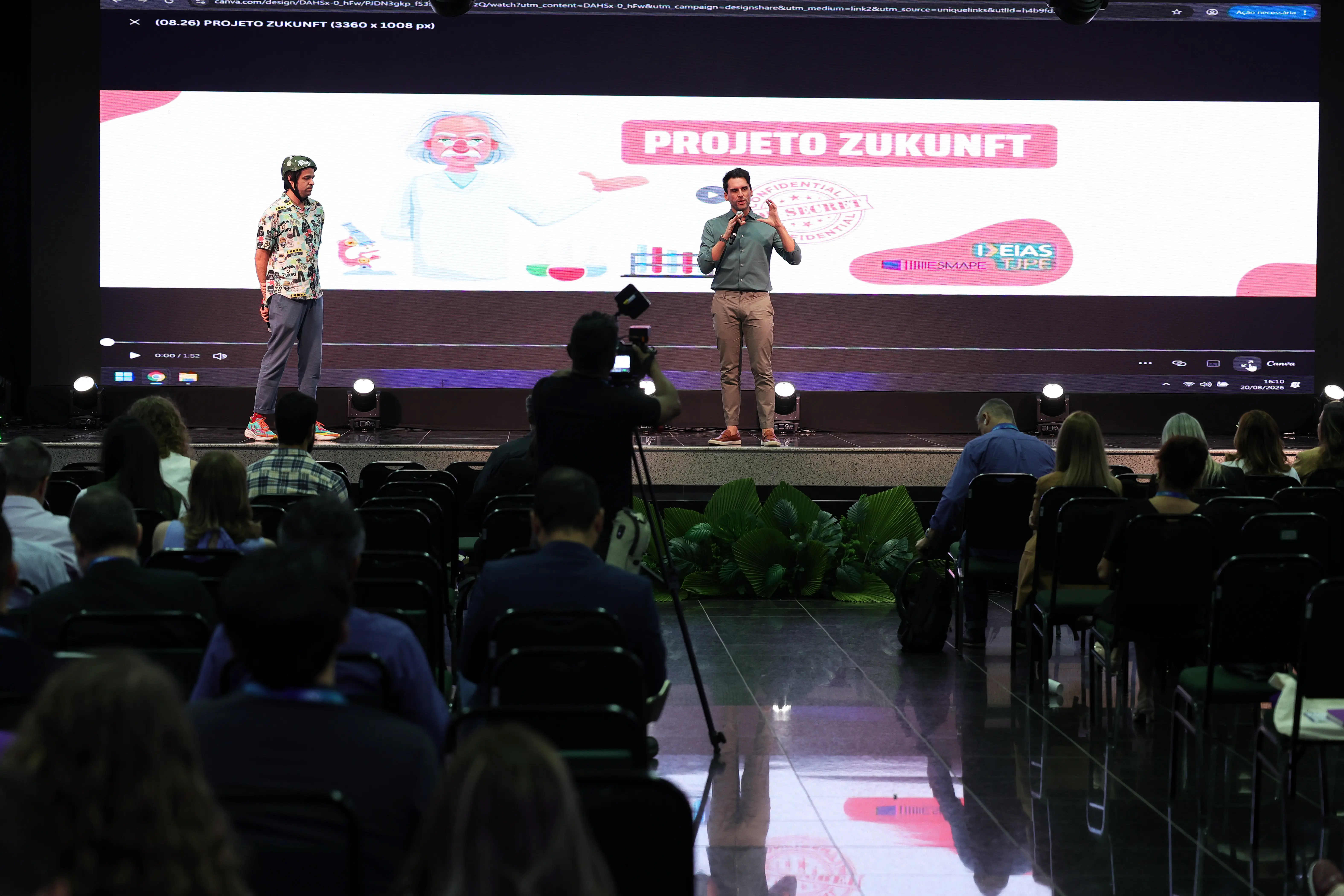 Uma platéia assiste a dois homens palestrando em um palco com um telão que exibe a frase "PROJETO ZUKUNFT" e o desenho de um cientista. Um cinegrafista filma a apresentação.