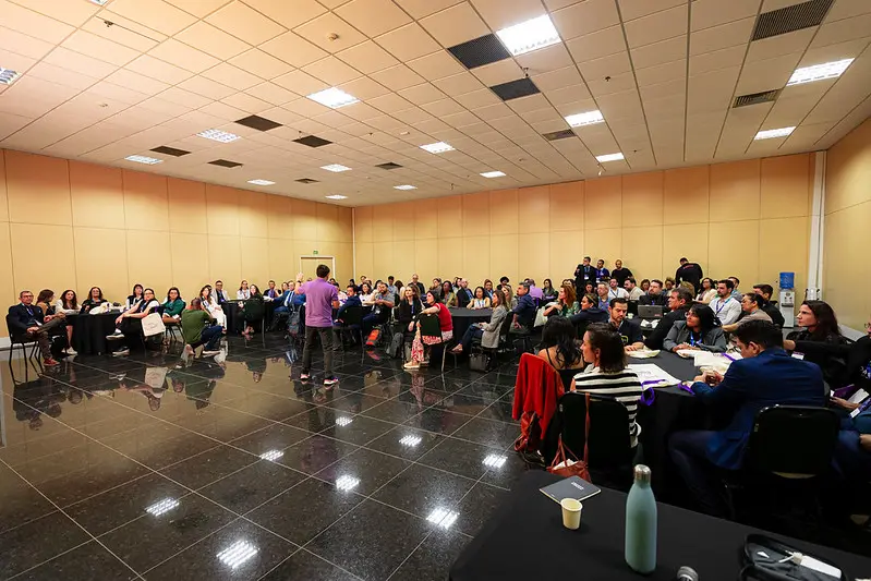 Foto horizontal em plano aberto que mostra uma grande sala com dezenas de pessoas sentadas em volta de mesas redondas, durante oficina no FestLabs Cuiabá. Á frente, em pé, de costas pra foto, está André Tamura conduzindo a atividade.