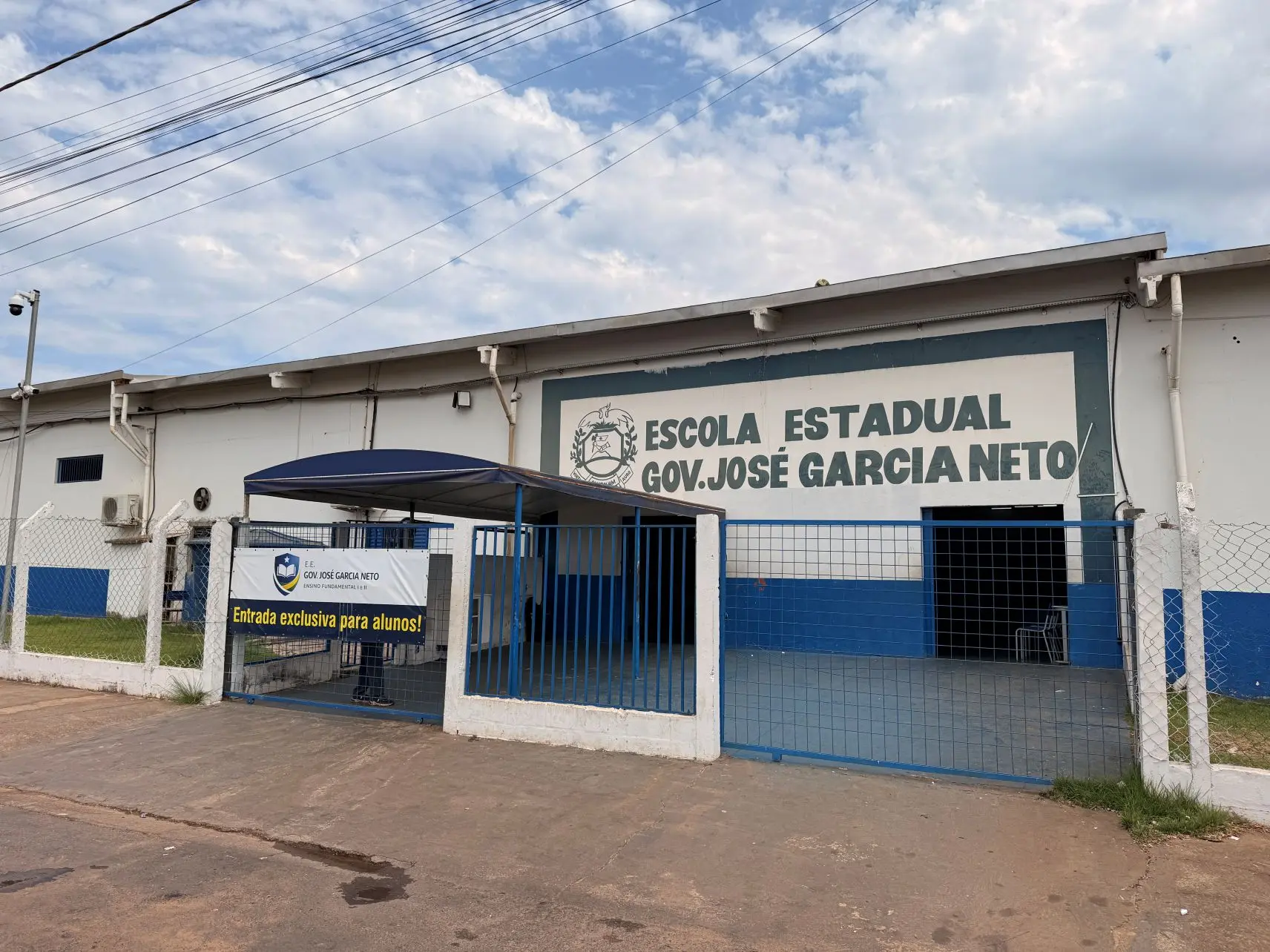 Foto horizontal em plano aberto que mostra a fachada da Escola Estadual Governador José Garcia Neto, em Várzea Grande. A escola é pintada de branco e azul, tem grade azul e um toldo azul entre o portão e a porta de entrada. O céu aparece azul ensolarado, com nuvens brancas.