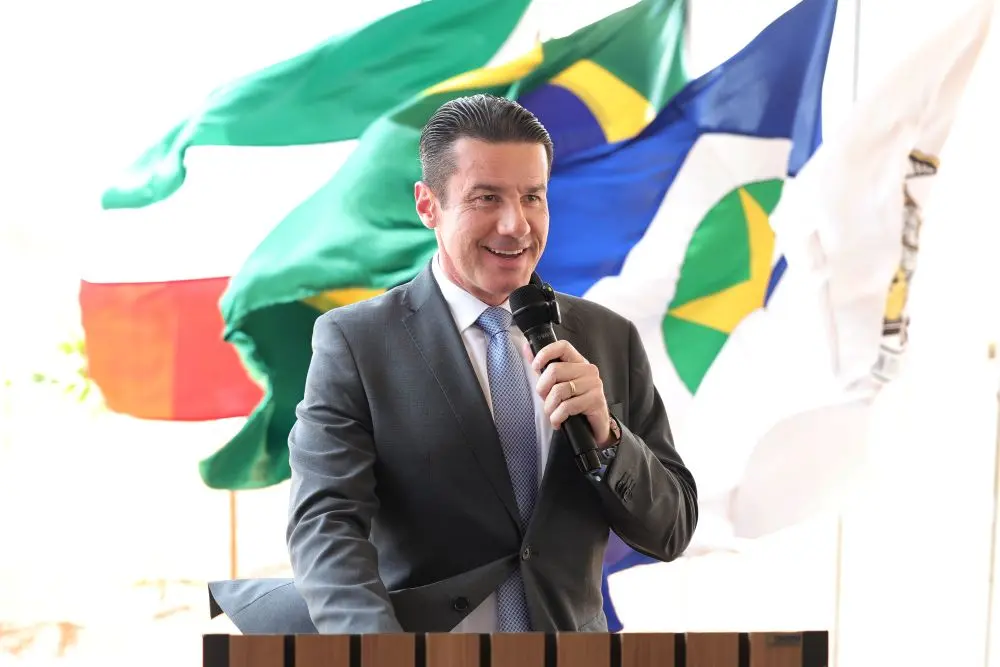 Foto horizontal que mostra o desembargador Rodrigo Curvo, da cintura pra cima, falando ao microfone em um púlpito e sorrindo. Ele é um homem branco, alto, magro, de cabelo preto, usando camisa branca, gravata azul clara, terno cinza. Atrás dele, aparecem mastros com bandeiras.