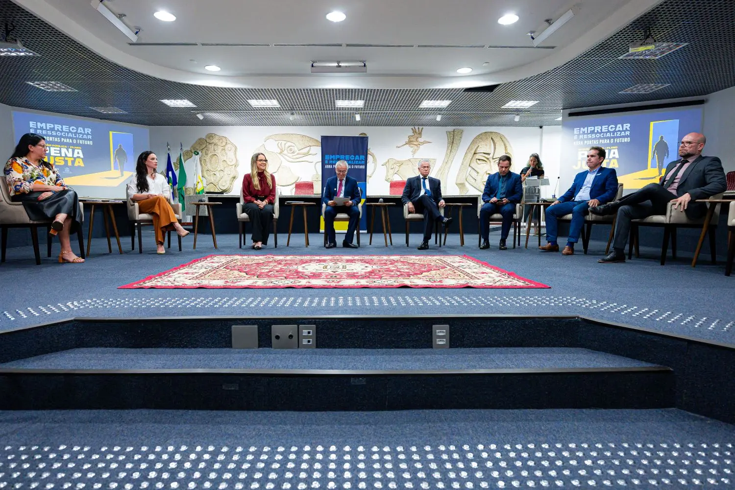 Foto horizontal em plano aberto que mostra oito pessoas sentadas em poltronas, no centro do plenário do Tribunal Regional do Trabalho de Mato Grosso.