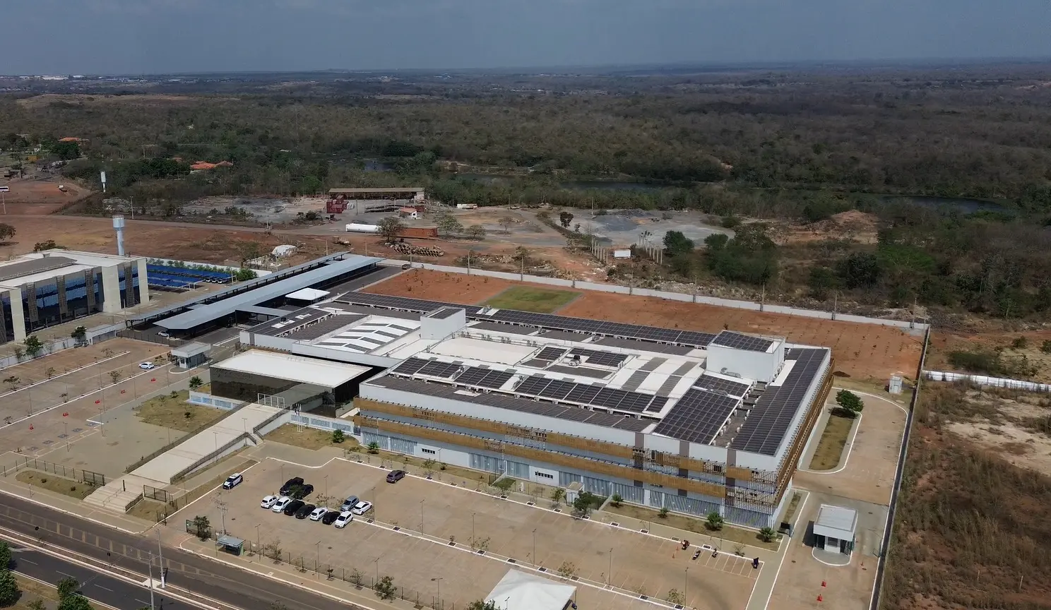 Imagem aérea que mostra o fórum de Várzea Grande e todo o seu terreno, tanto da frente e lateral, onde é o estacionamento, quanto dos fundos, onde estão 1.500 mudas de árvores plantadas. Atrás do terreno do fórum, aparece ainda uma extensa área de vegetação e o céu ensolarado.