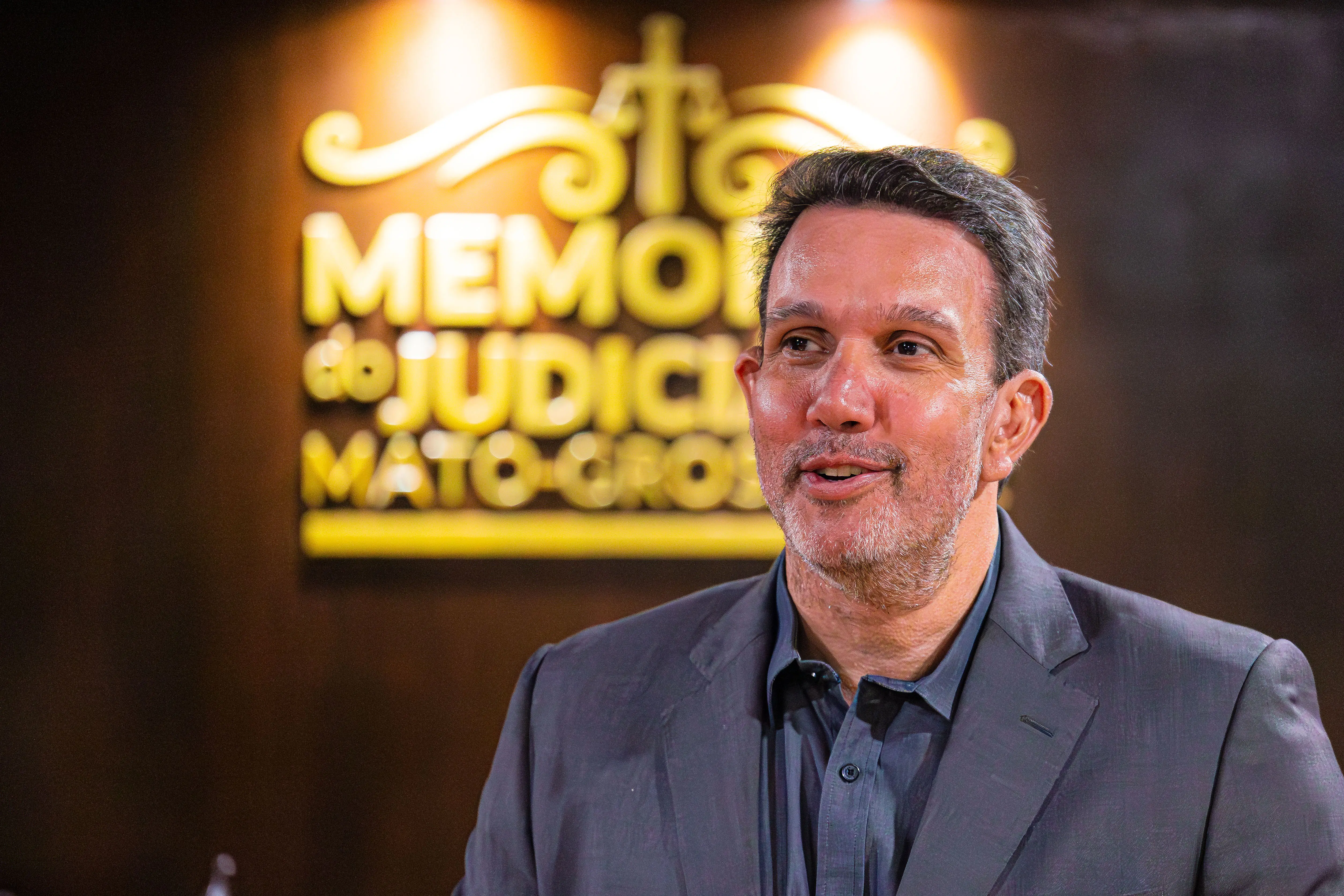 Homem de terno escuro e barba rala sorri olhando para o lado. Ao fundo, letreiro dourado na parede exibe "MEMÓRIA do JUDICIÁRIO MATO-GROSSENSE" sob o símbolo da justiça.