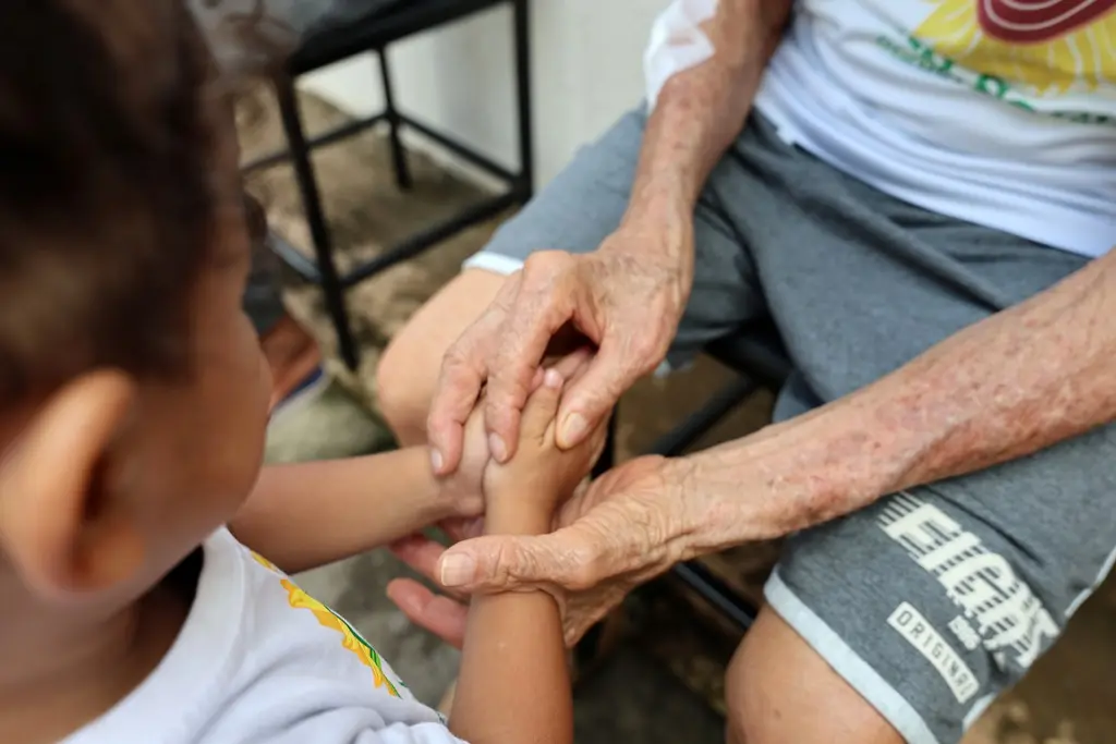 Foto horizontal que mostra detalhe das mãos de uma criança pedindo bênção a um idoso.