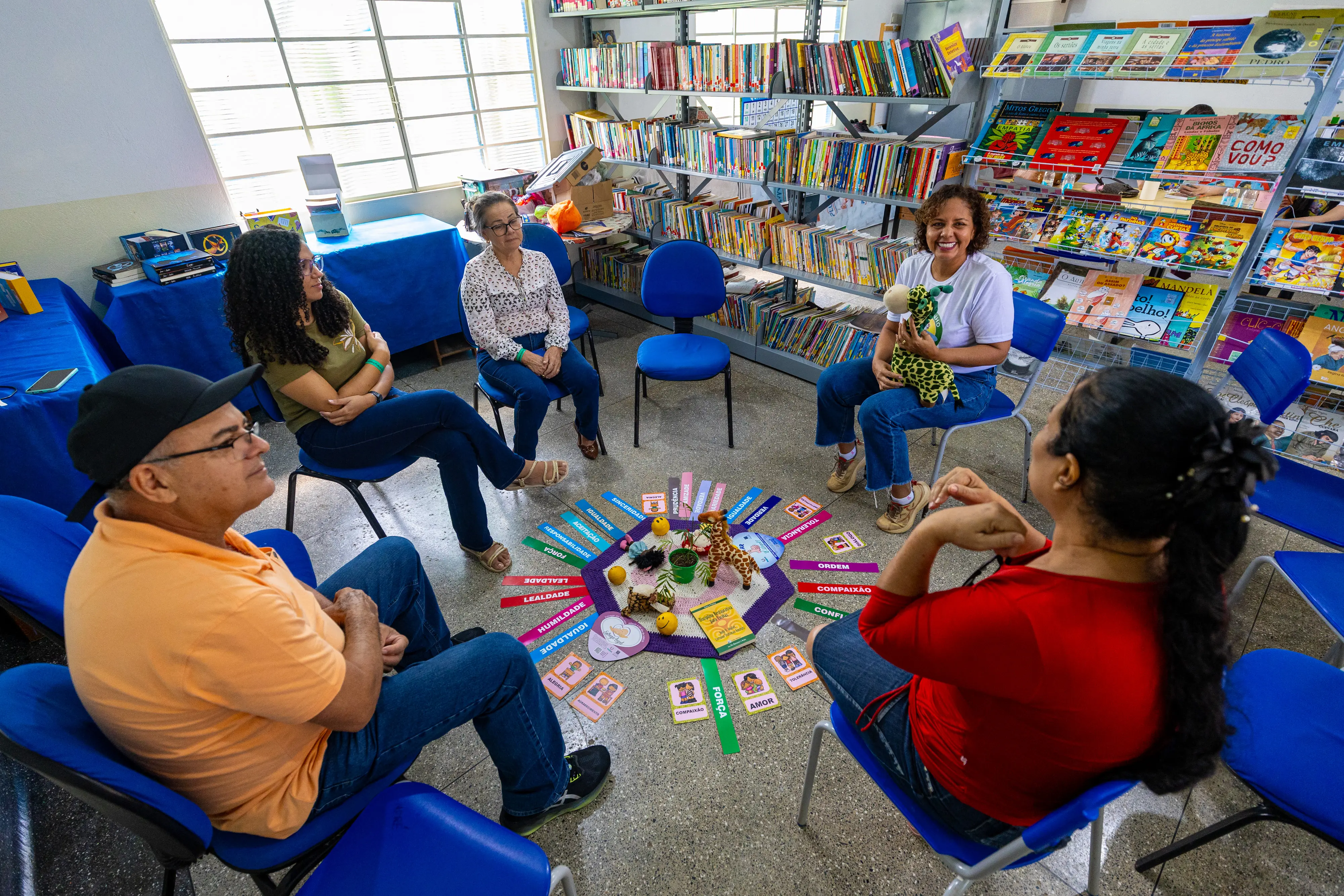 Adultos formam uma roda de conversa em uma biblioteca. No centro, um tapete com palavras e brinquedos. Uma mulher segura uma girafa de pelúcia. Ao fundo, estantes repletas de livros coloridos.