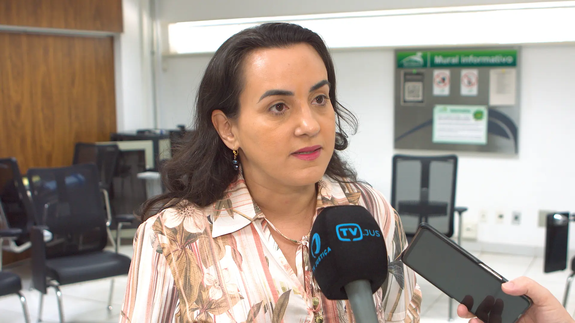 Mulher de pele clara, cabelos escuros soltos, veste roupa clara e fala ao microfone com logomarca de emissora. Está em ambiente interno institucional, com mesa e computador ao fundo.