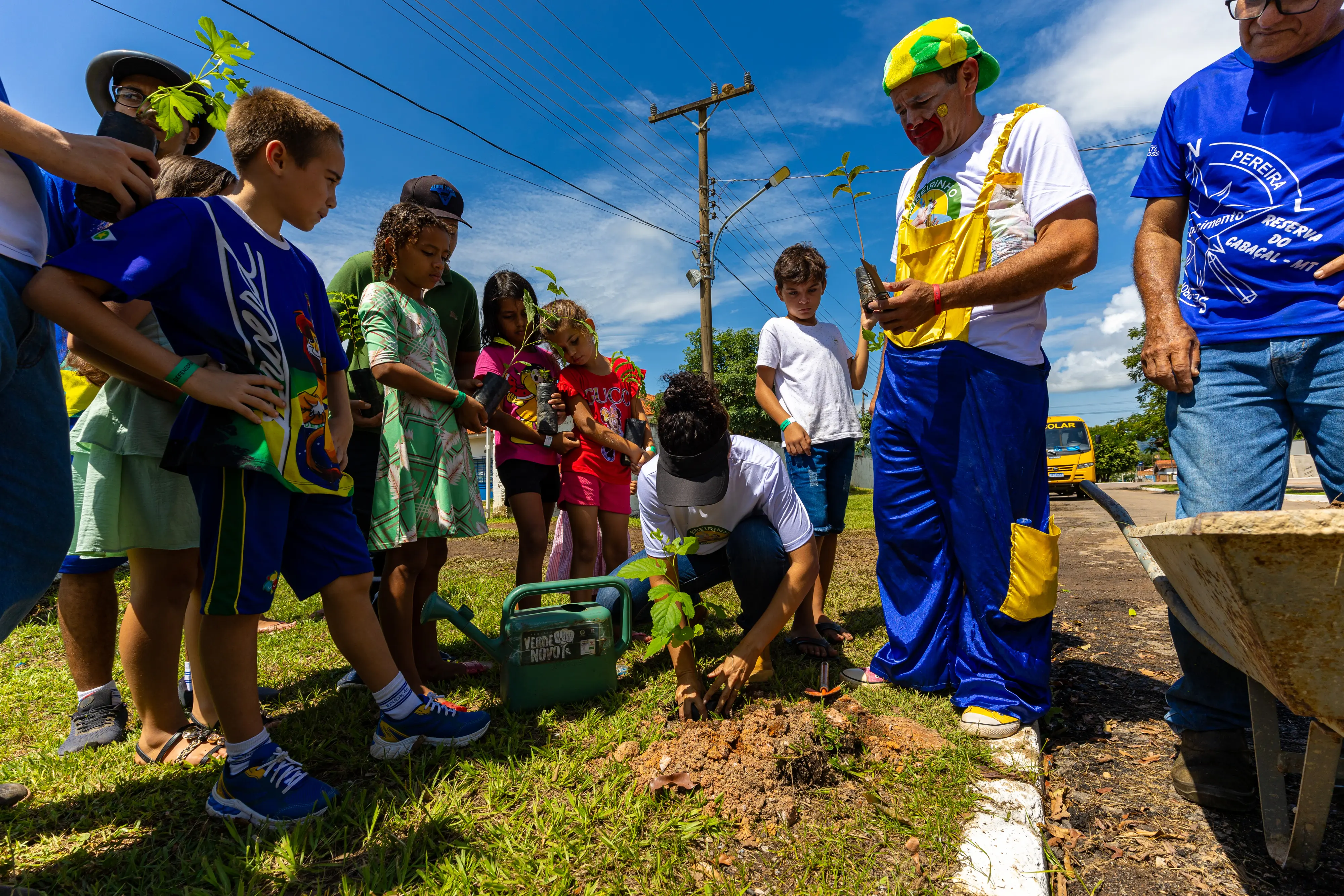 Ao ar livre, crianças e um palhaço observam uma mulher plantando uma muda no gramado. Há um regador verde e um carrinho de mão. O dia é ensolarado sob céu azul na Reserva do Cabaçal.