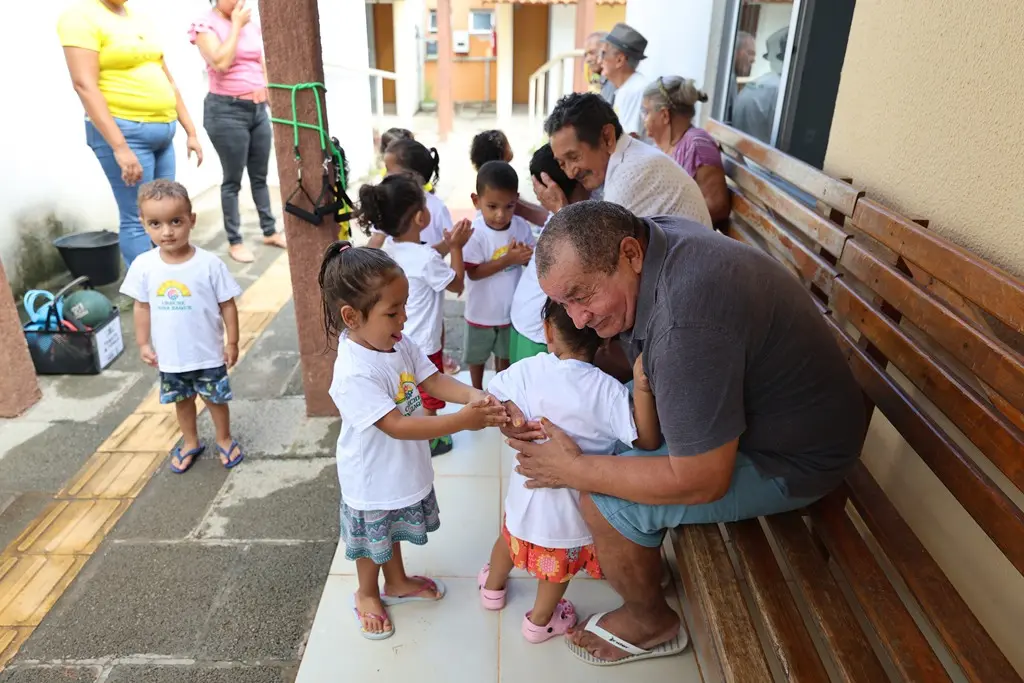 Foto horizontal colorida que mostra um grupo de crianças da creche Nina Zaque visitando idosos na Vila de Luz. Alguns deles dão abraços, outros pedem bênção aos idosos, que estão sentados em bancos de madeira em frente à casa. 