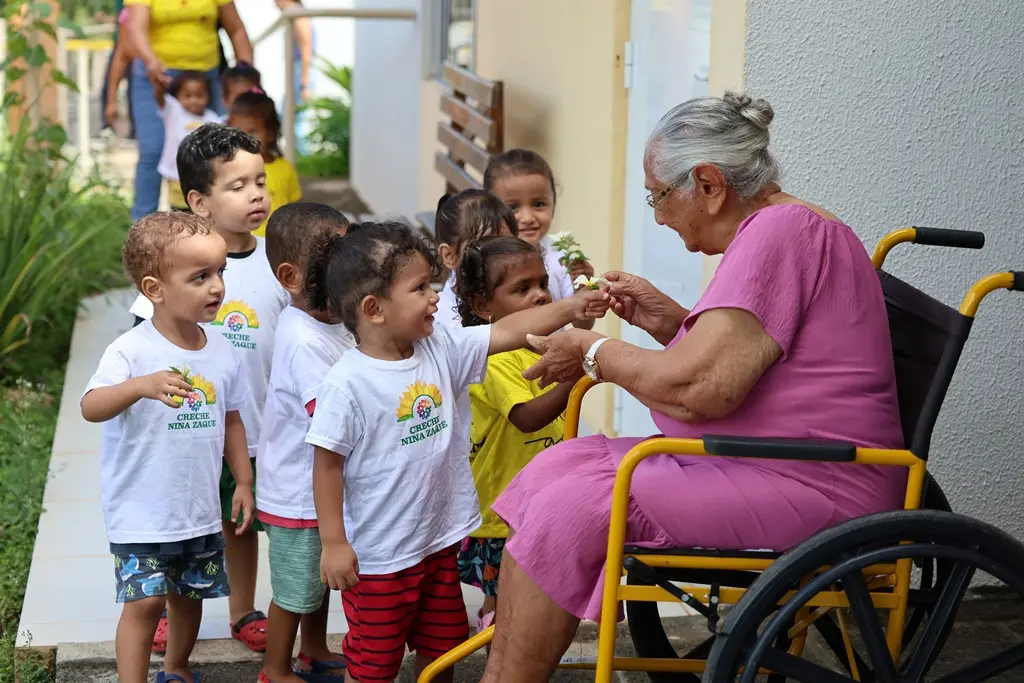 Foto horizontal colorida que mostra um grupo de crianças da creche Nina Zaque visitando a idosa Maria, na Vila de Luz. Um menininho entrega a ela flores.