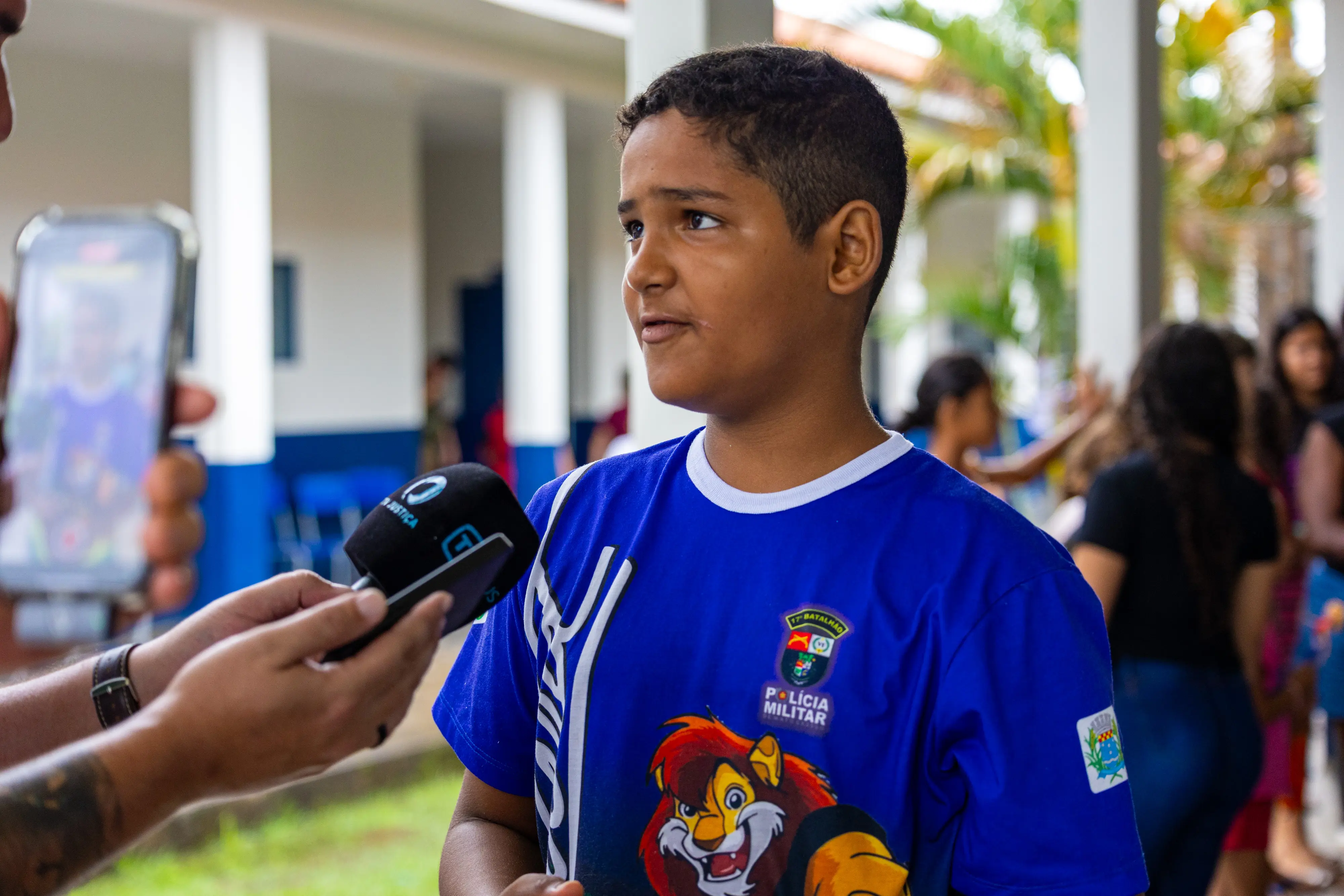 Menino pardo de camiseta azul com estampa do Proerd e brasão da Polícia Militar concede entrevista. Um microfone da TV Justiça e um celular apontam para ele. Ao fundo, um corredor de escola.