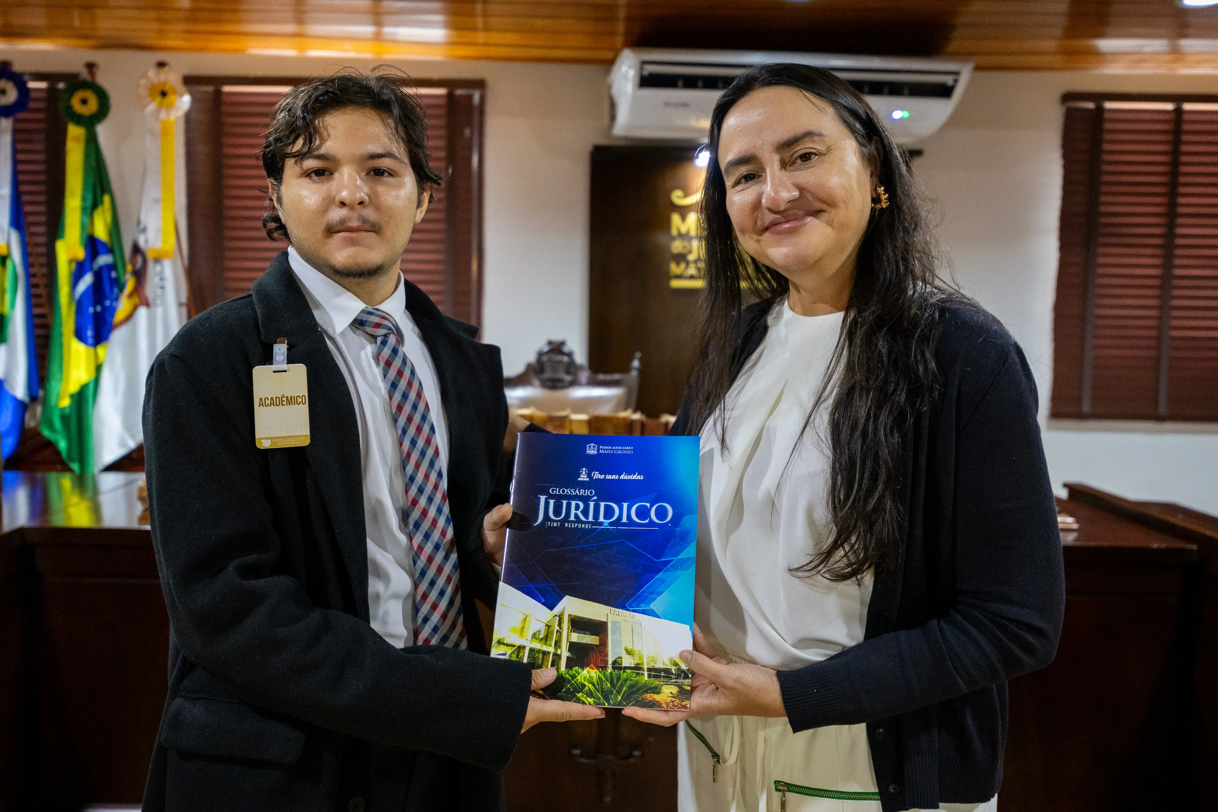 Um jovem de terno preto e gravata xadrez e uma mulher de cabelos longos escuros seguram juntos um exemplar do Glossário Jurídico do TJMT. Ao fundo, bandeiras do Brasil e de Mato Grosso.