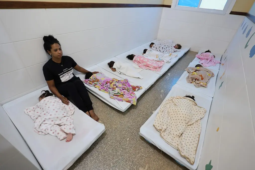 Foto horizontal colorida que mostra o dormitório da creche Nina Zaque com 10 crianças dormindo nas caminhas e uma professora sentada. 