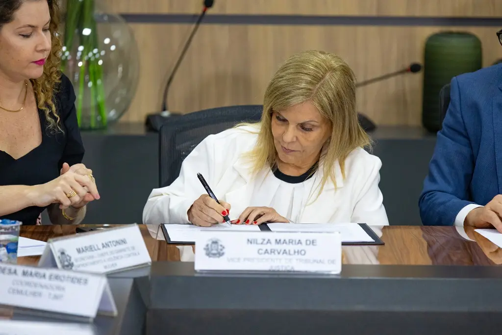 Foto horizontal que mostra a vice-presidente do TJMT, desembargadora Nilza Maria Pôssas de Carvalho, sentada, assinando um documento. Ela é uma senhora de pele clara, loira, usando vestido e blazer brancos.