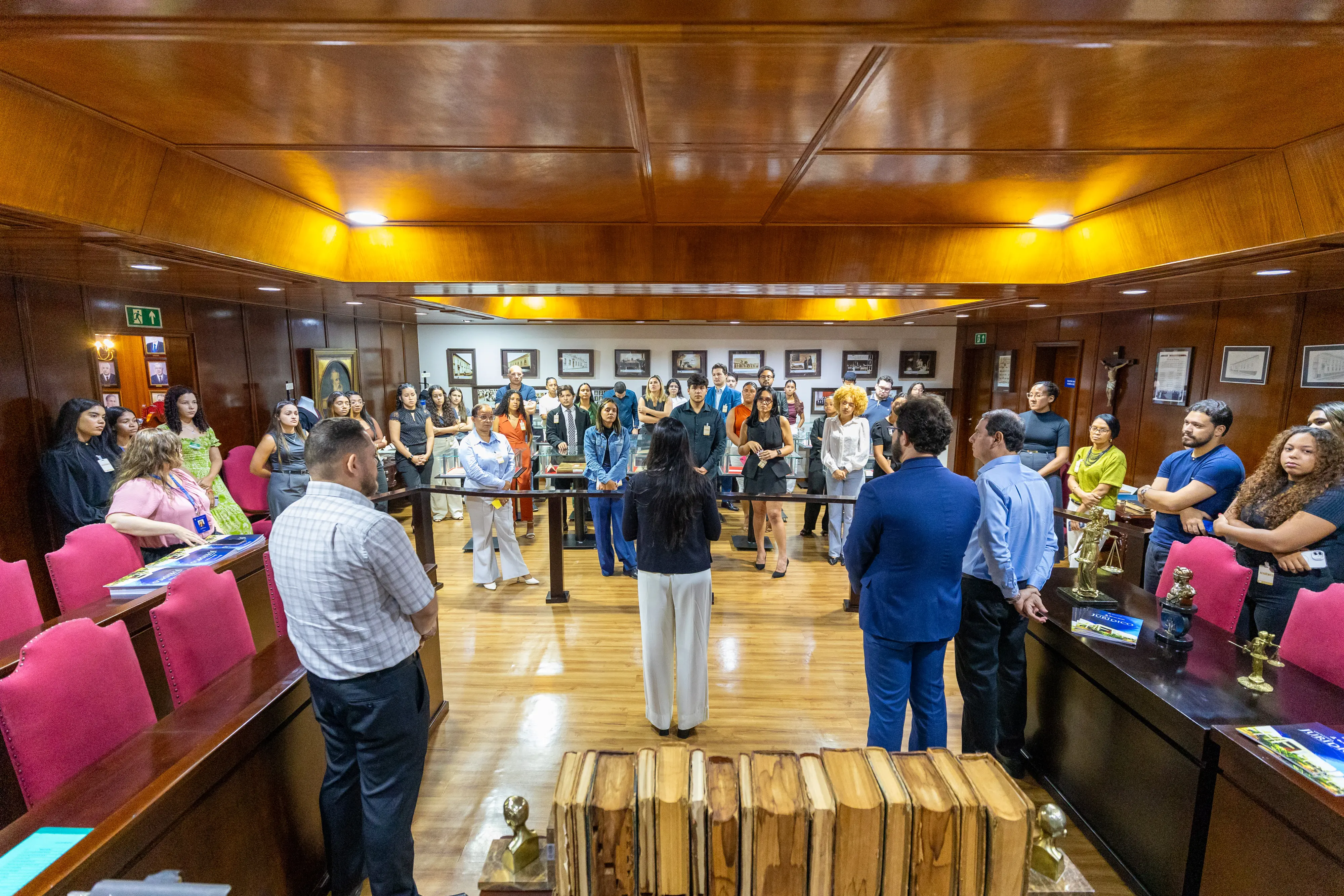 Grupo de pessoas reunidas em uma sala histórica de madeira nobre, com retratos nas paredes, observa uma apresentação conduzida por uma mulher ao centro. Sobre as mesas, exemplares do Glossário Jurídico.
