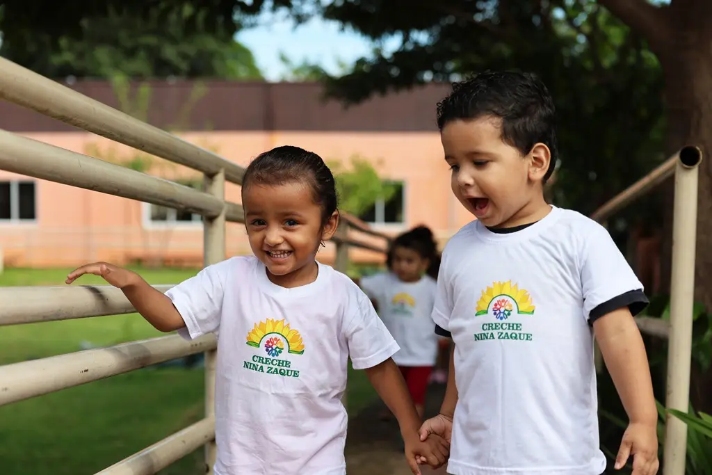 Foto horizontal colorida que mostra uma menininha e um menininho de cerca de 3 anos de idade, andando de mãos dadas em uma rampa e sorrindo. Eles estão usando camiseta branca com estampa da creche Nina Zaque. Ao fundo, o local é um gramado com árvores e plantas e há mais crianças andando em fila.
