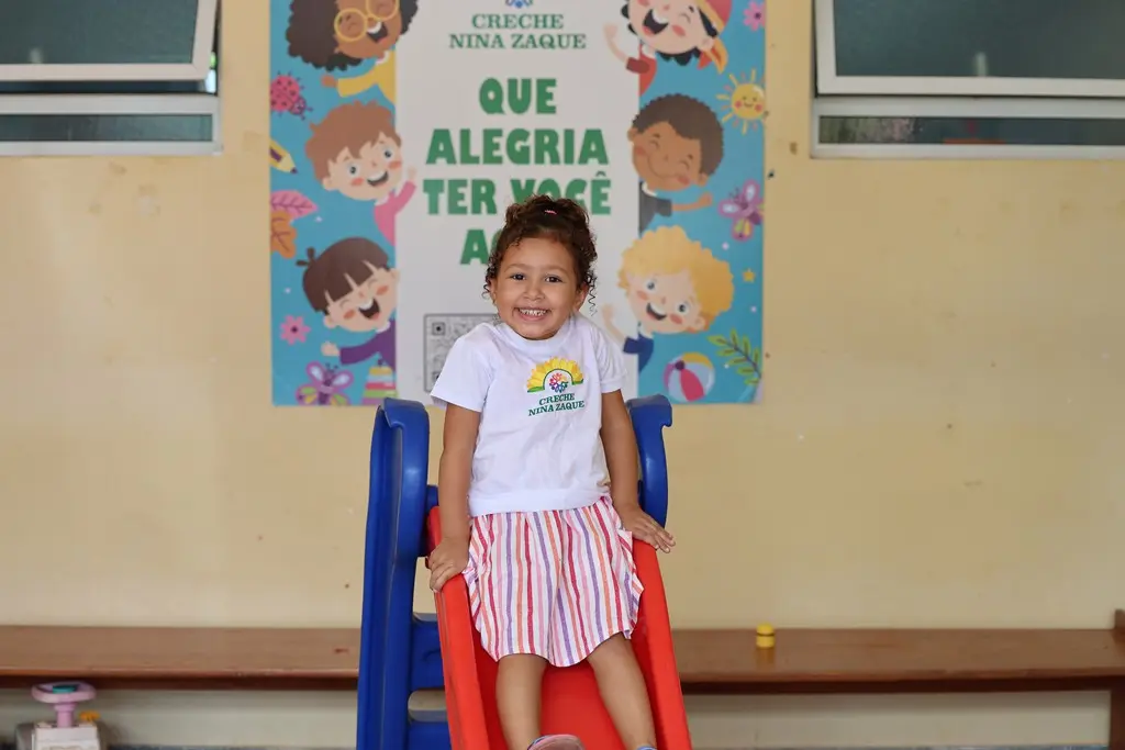 Foto horizontal colorida que mostra uma menina de 3 anos de idade com a camiseta da creche Nina Zaque brincando no escorregador, no pátio da creche.